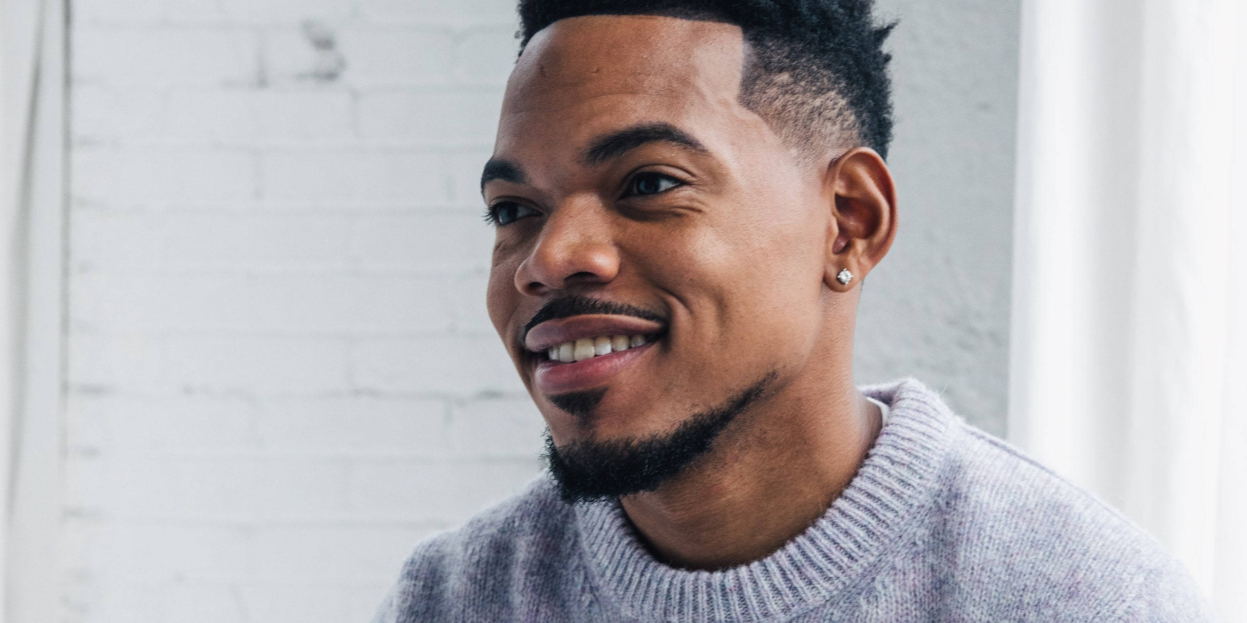 帥氣的Chance the Rapper 壁紙 — 一位留鬍鬚、戴耳環的微笑男士，身穿灰色毛衣，背景是白色磚牆。