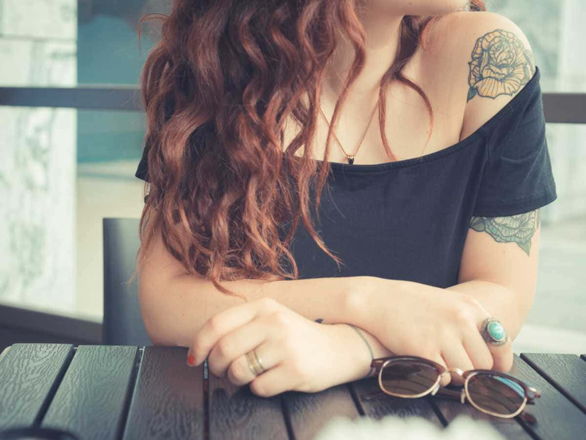 帥氣紋身女孩 壁紙 — 一位擁有捲曲赤褐色頭髮和玫瑰紋身的女士，身穿黑色露肩上衣，將手臂擱在木桌上。
