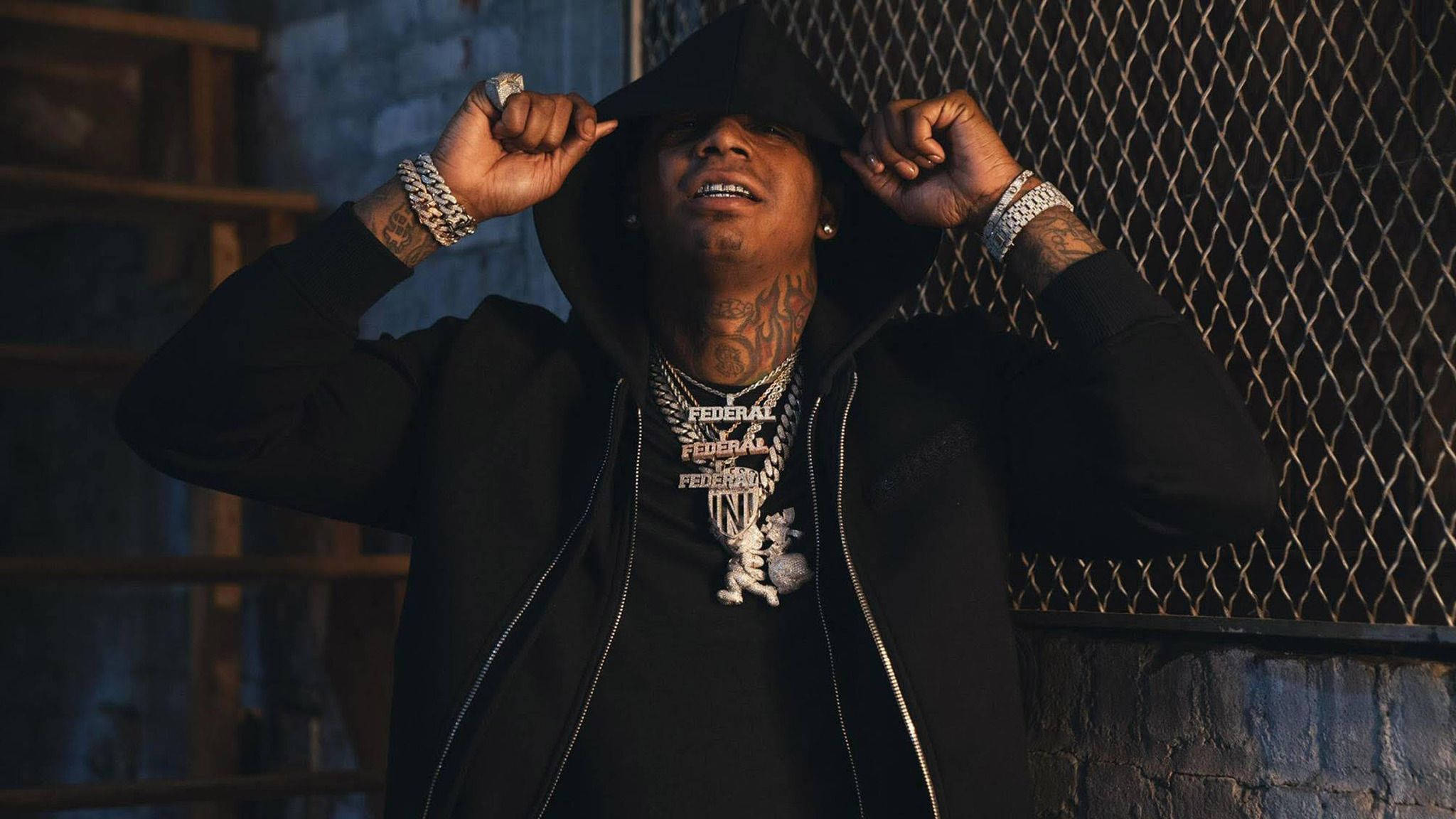 帥氣饒舌歌手 Moneybagg Yo 壁紙 — 一個有紋身和鏈條的男人在昏暗的城市燈光下拉起連帽衫的帽子，靠近鐵絲網圍欄。