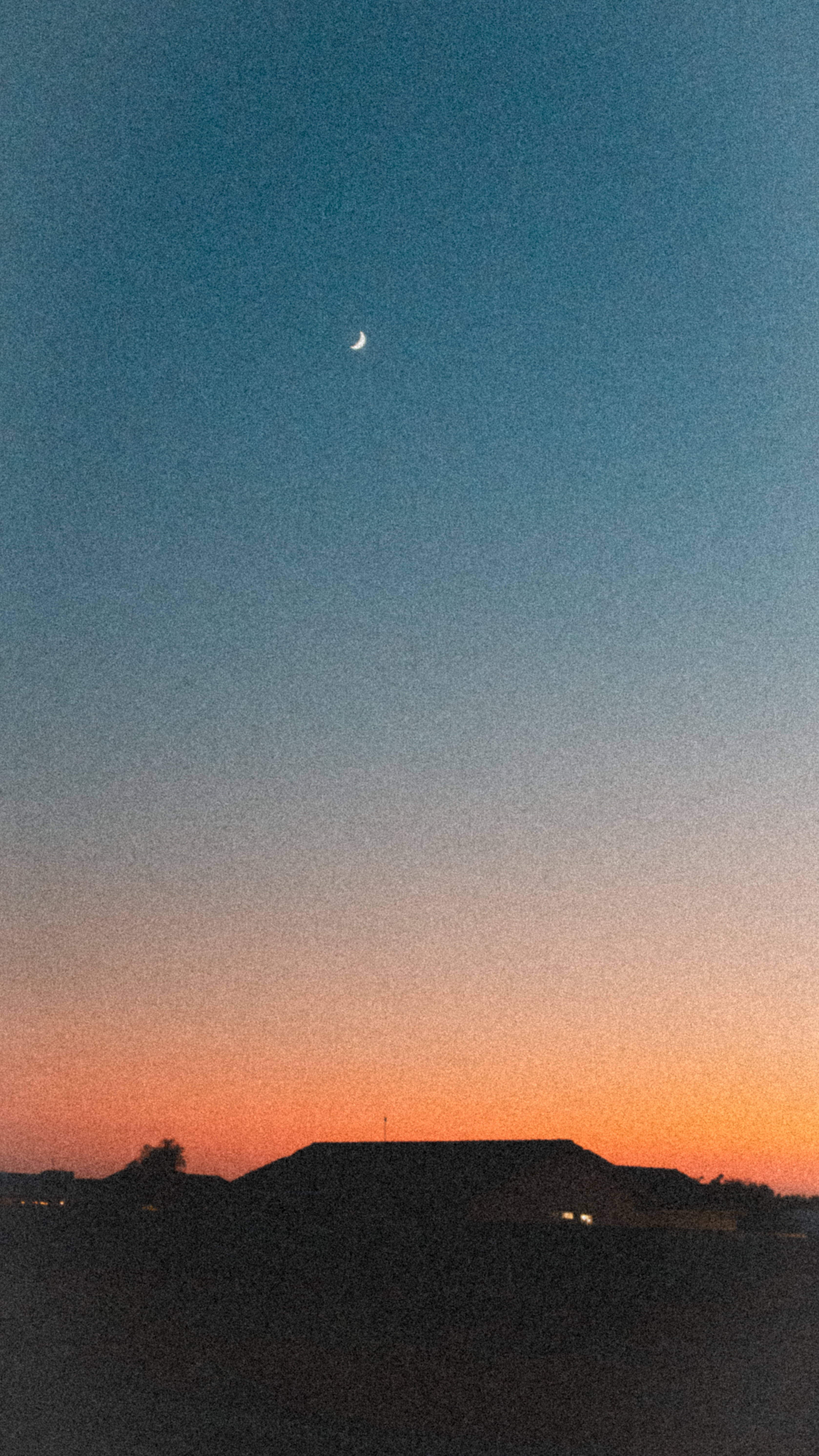 帶房屋剪影的美學日落 壁紙 — 一彎新月在從深藍色過渡到溫暖橙色的暮色天空中發光，映襯著屋頂的黑暗輪廓。