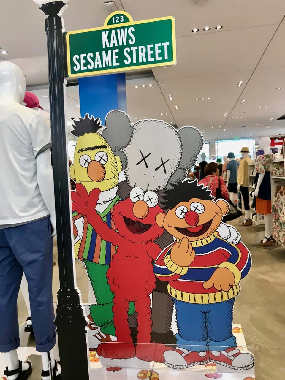Kaws Sesame Street 桌布 壁紙 — 帶有KAWS X眼睛的芝麻街角色站在零售店的街道標誌下，店內有模特兒和購物者。