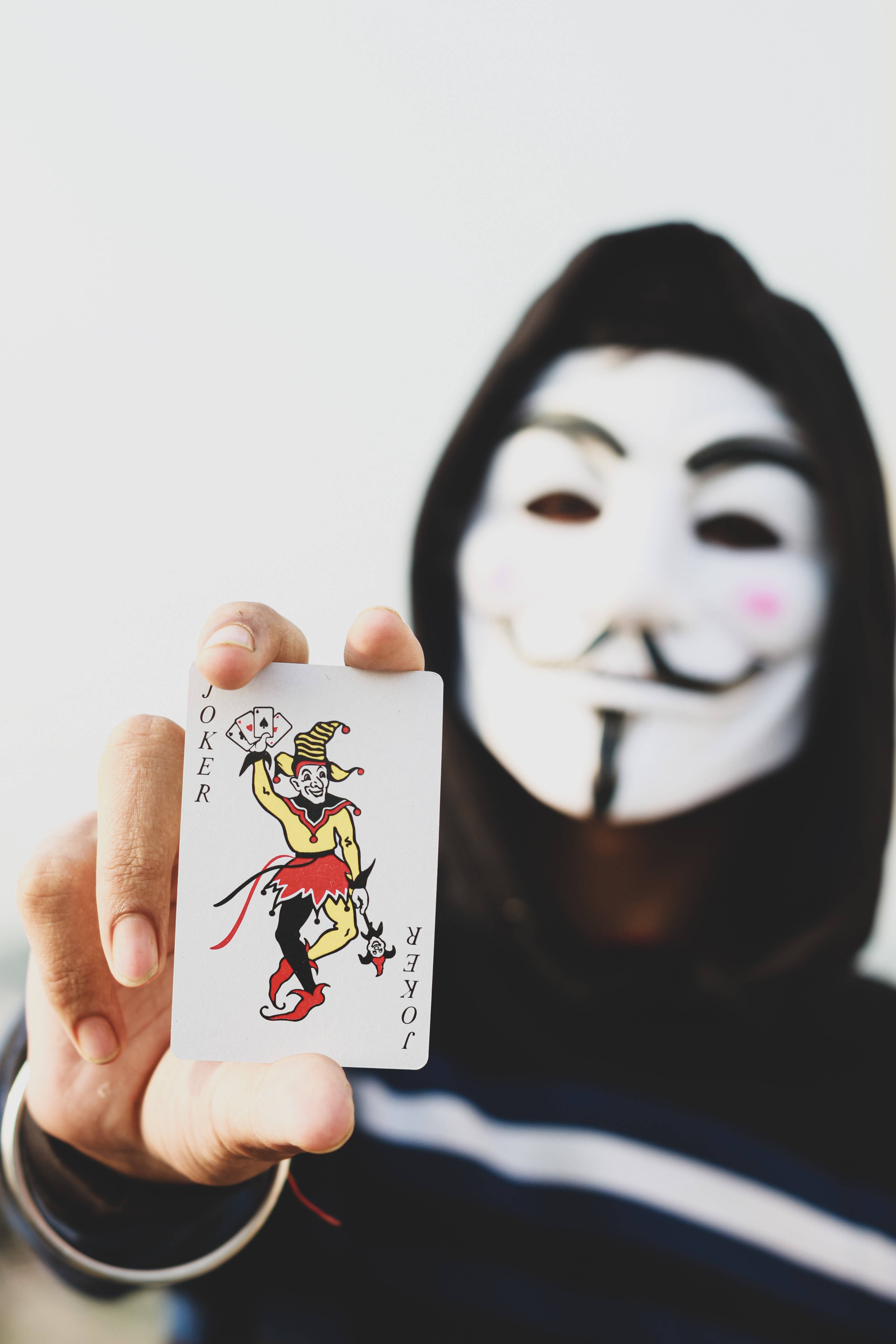 帶有撲克牌的神秘黑客面具——網路反抗的象徵 壁紙 — 一個戴著蓋伊·福克斯面具的人，在純白色背景前舉起一張小丑撲克牌。