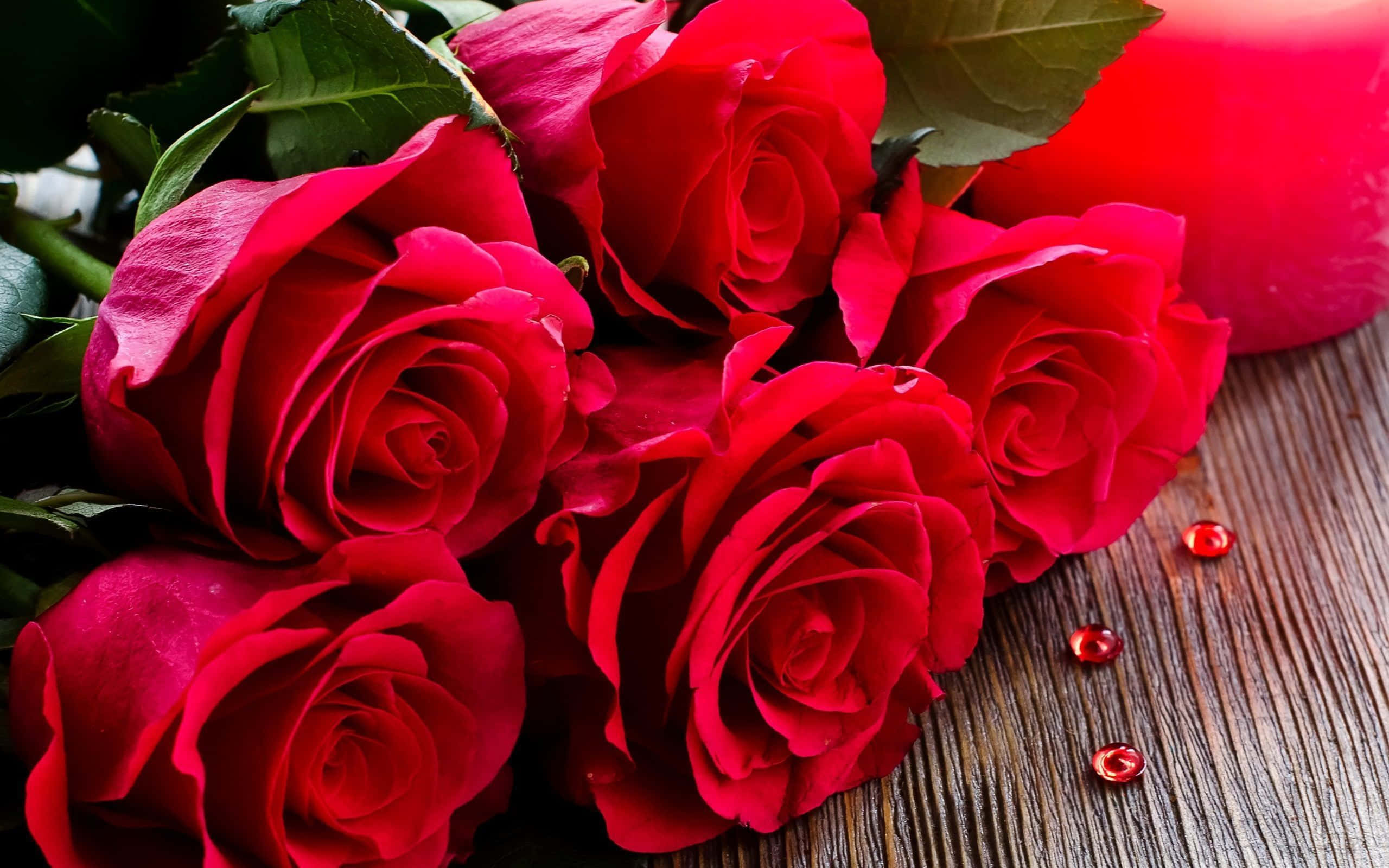 Red Rose 桌布 壁紙 — 帶有露珠的鮮豔紅玫瑰放在質樸的木質表面上，旁邊有一根發光的紅色蠟燭。