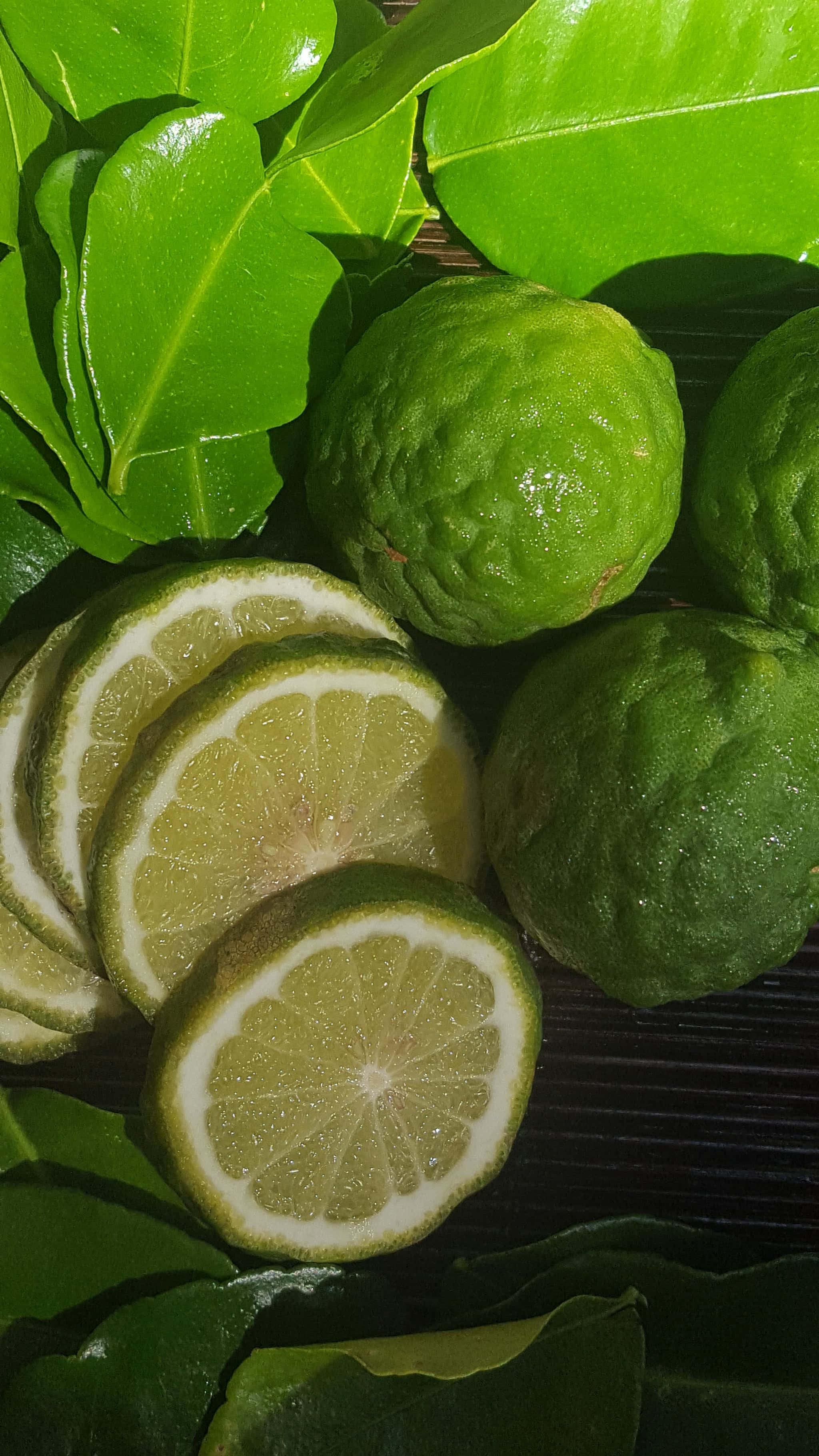 帶綠葉的檸檬葉水果 壁紙 — 新鮮青檸切片和完整青檸，依偎在光澤的熱帶葉子中，放在深色木質表面上。