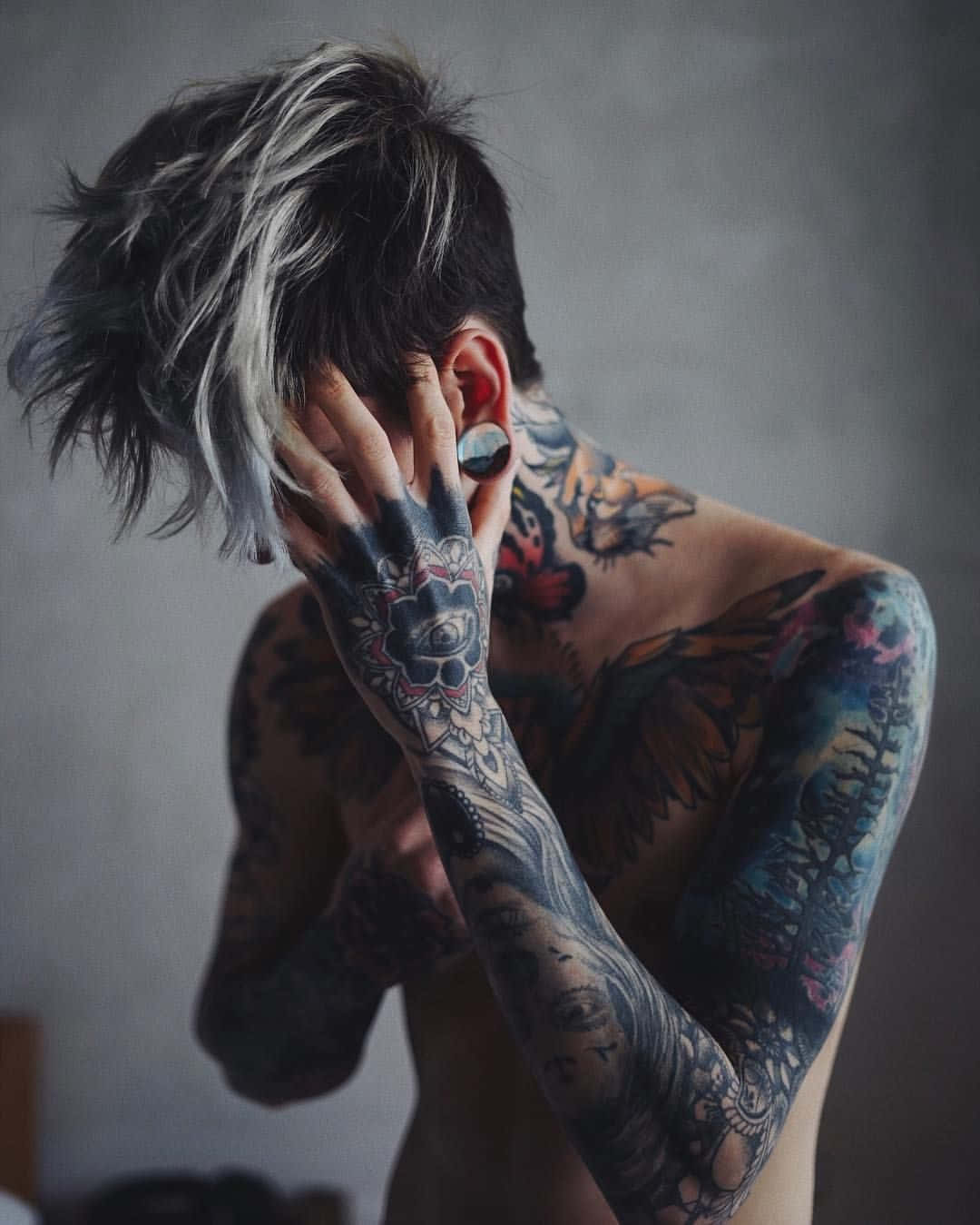 帶著誘惑眼神的紋身男孩 壁紙 — 一位有紋身的人，銀色和黑色頭髮，用有紋身的手遮住臉，背景是柔和的灰色。