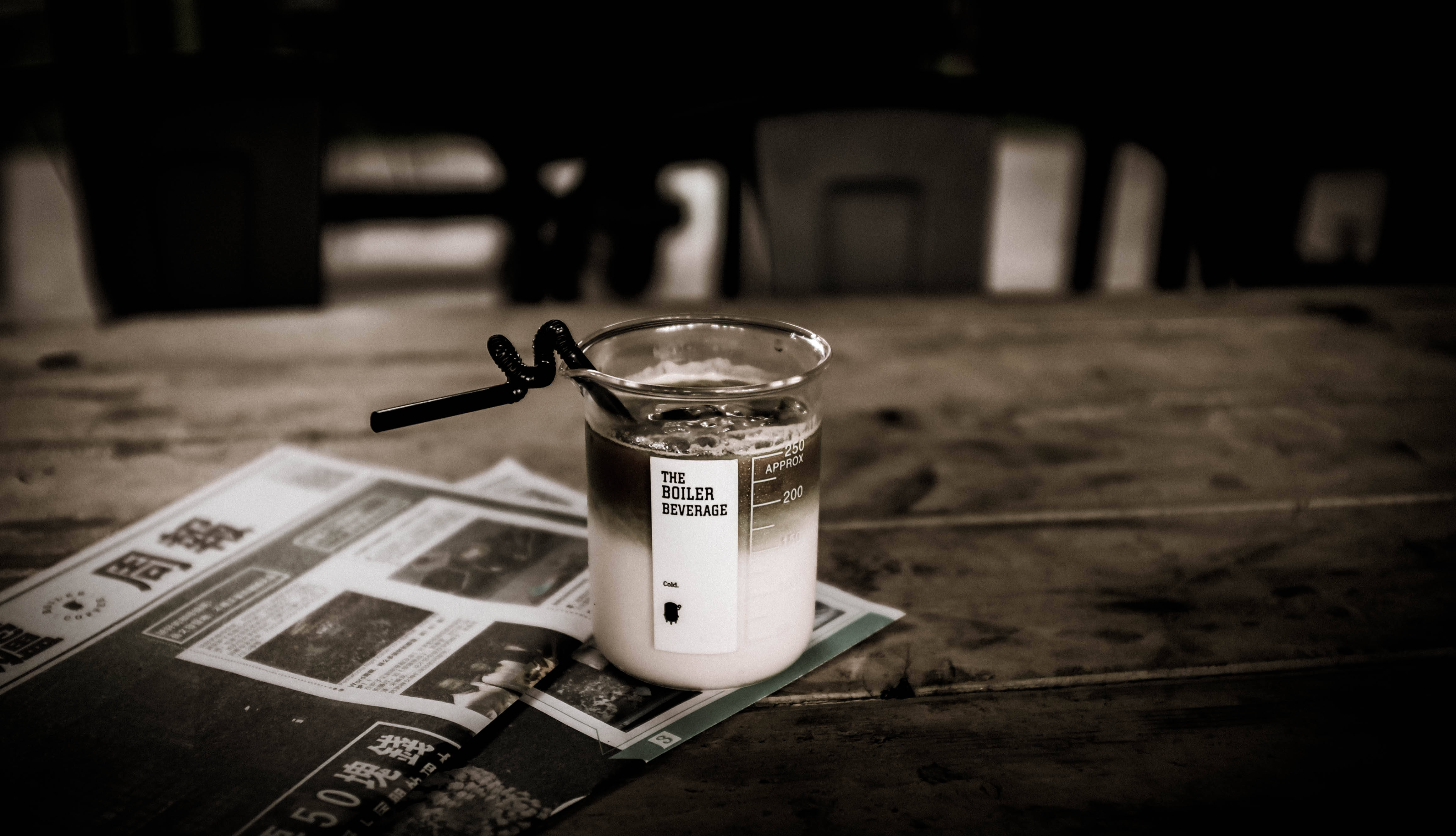 帶黑色吸管的燒杯 壁紙 — 一個懷舊色調的燒杯，標有「THE BOILER BEVERAGE」，帶有黑色吸管，放在一張舊木桌上的報紙上。