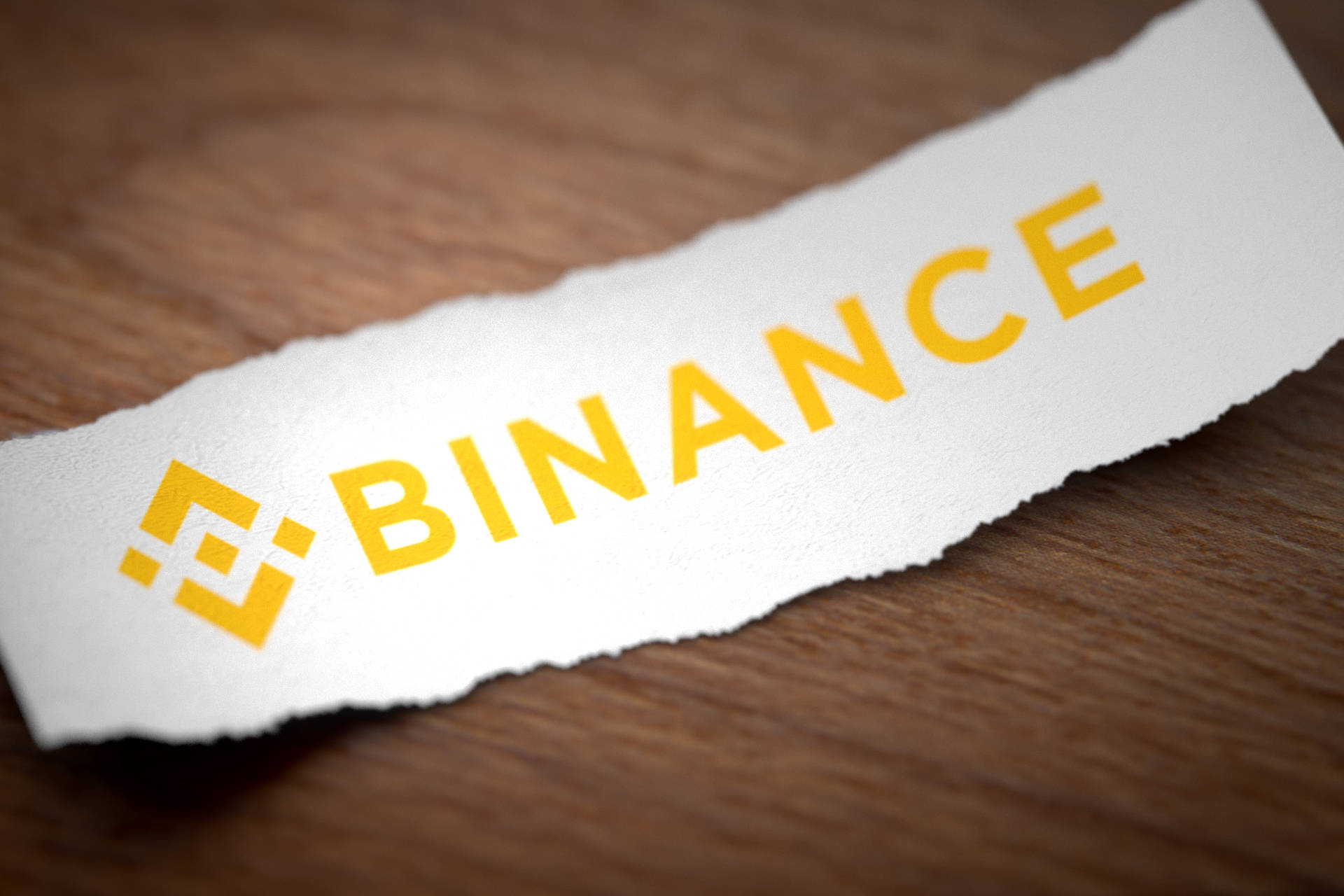 幣安撕裂紙張 壁紙 — 一條撕裂的白色紙條帶有黃色 Binance 標誌，放在溫暖的木質表面上，景深較淺。