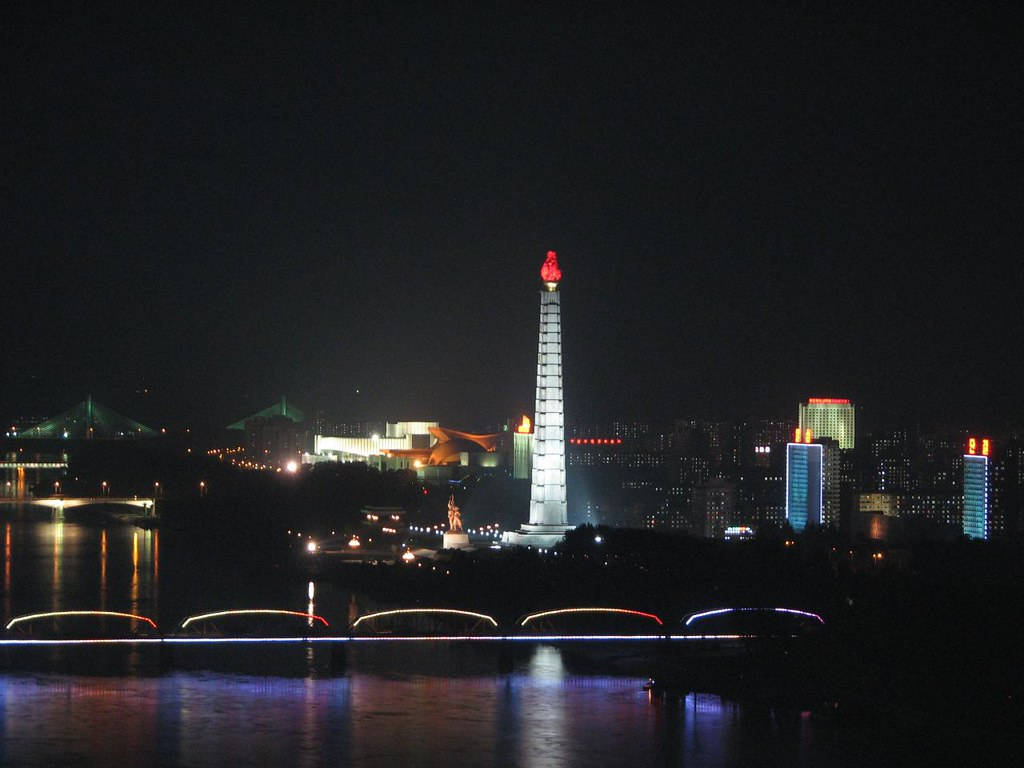 夜間景色，一座發光的紀念碑立於水上，背景有橋樑和城市燈光。