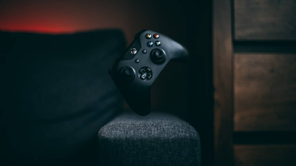 平衡的Xbox遊戲控制器 壁紙 — 一個黑色Xbox手掣放在灰色織物扶手上，背景為深色木材，帶有溫暖的環境燈光。