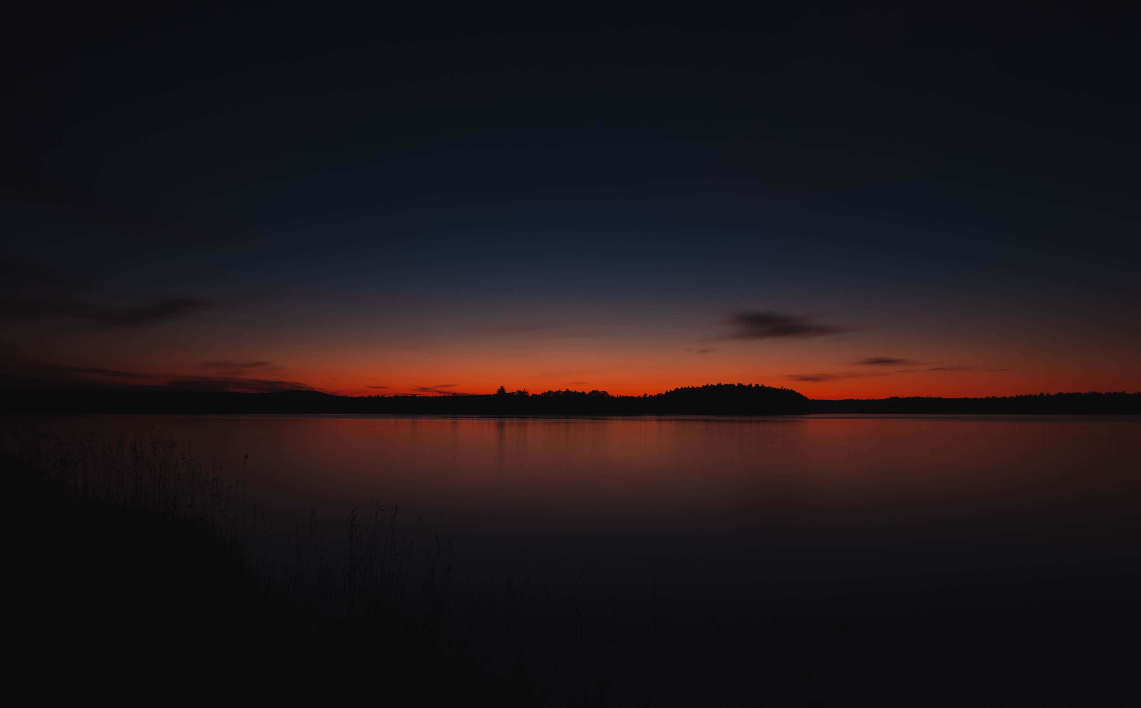 Red Sunset 桌布 壁紙 — 平靜的湖面倒映著熾熱的橙色日落，逐漸消失在深靛藍色的天空中，地平線上有樹木的剪影。