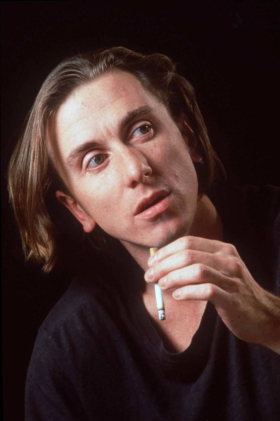 年輕 Tim Roth 長棕髮抽菸肖像 壁紙 — 一個留著及肩長髮的男人，在戲劇性的肖像照中，手持香煙，背景是純黑色。