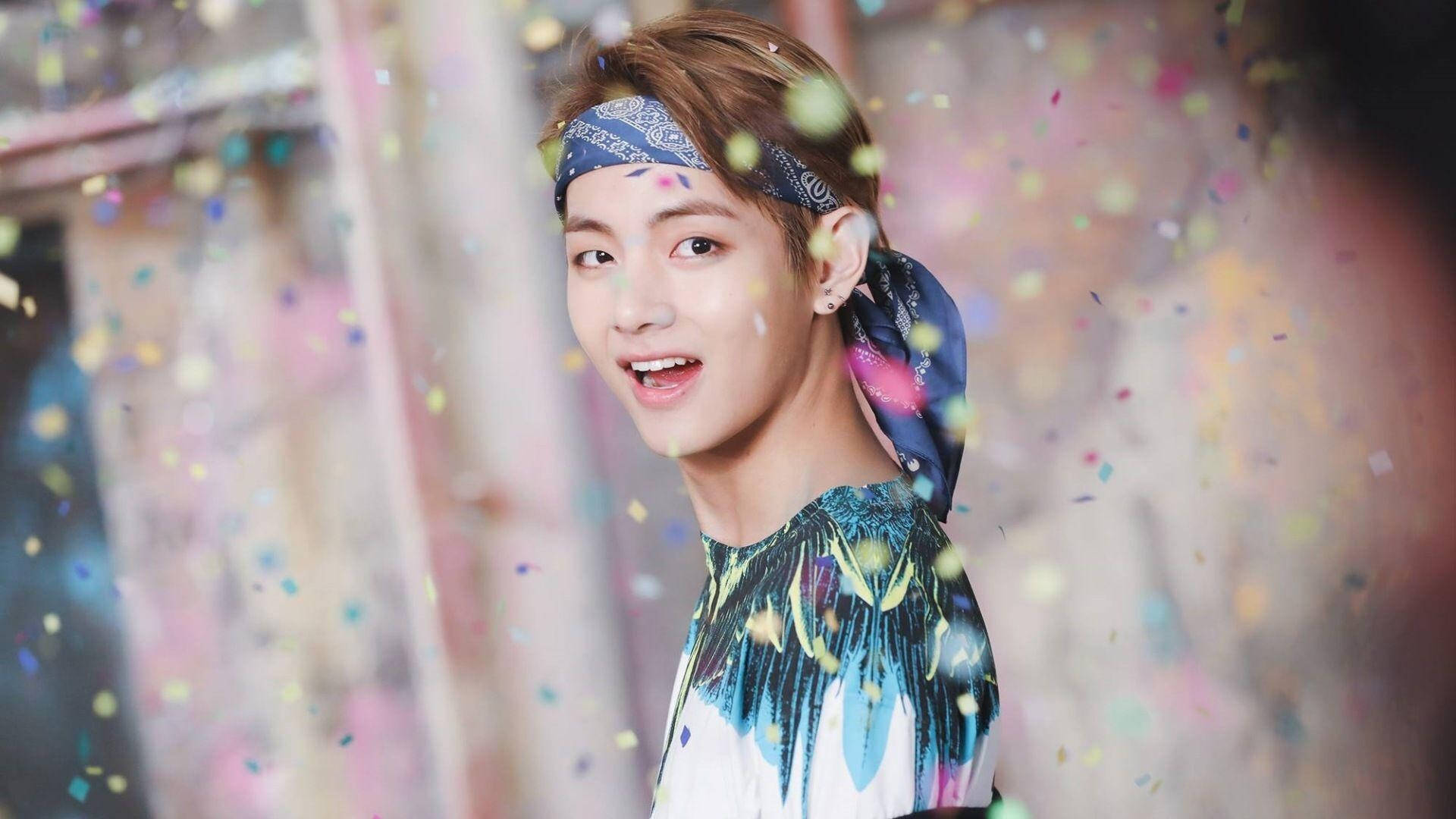 年輕BTS V 藍色頭巾 壁紙 — 一位戴著藍色圖案頭帶的年輕男子在充滿活力的攝影棚中微笑，彩色紙屑在他周圍飄落。