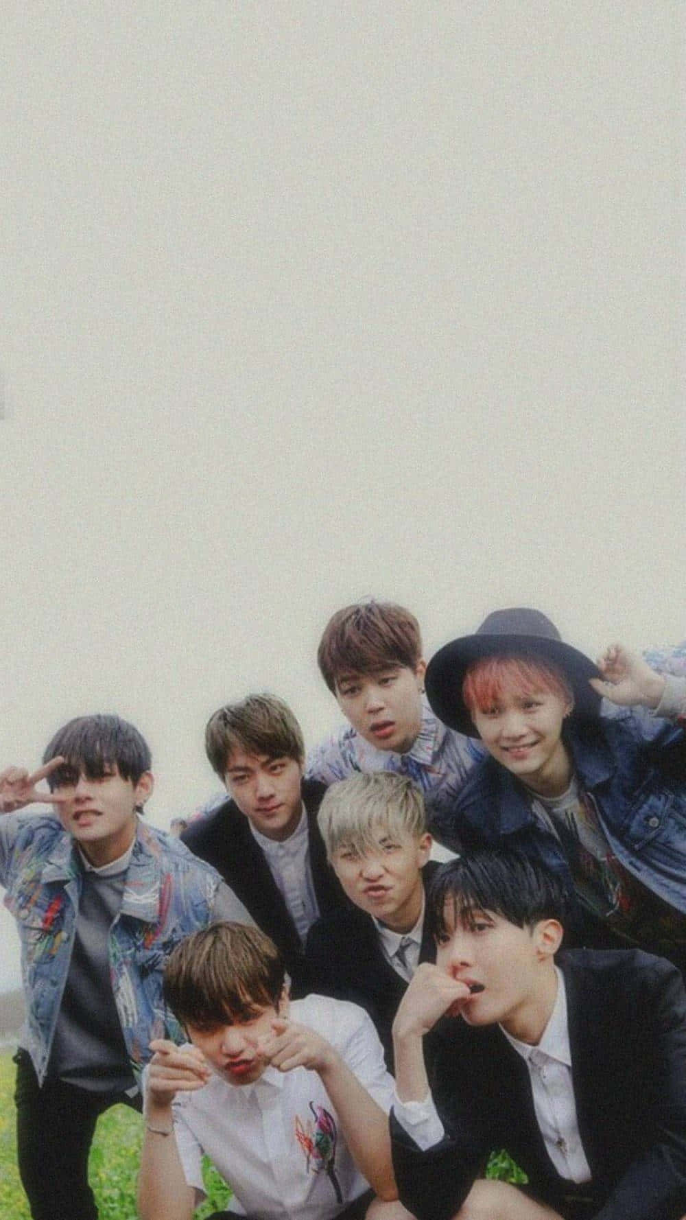 年輕BTS拍攝照 壁紙 — 七位年輕男子身穿休閒服裝，在草地上以多種表情和手勢，對著朦朧的天空擺出俏皮的姿勢。