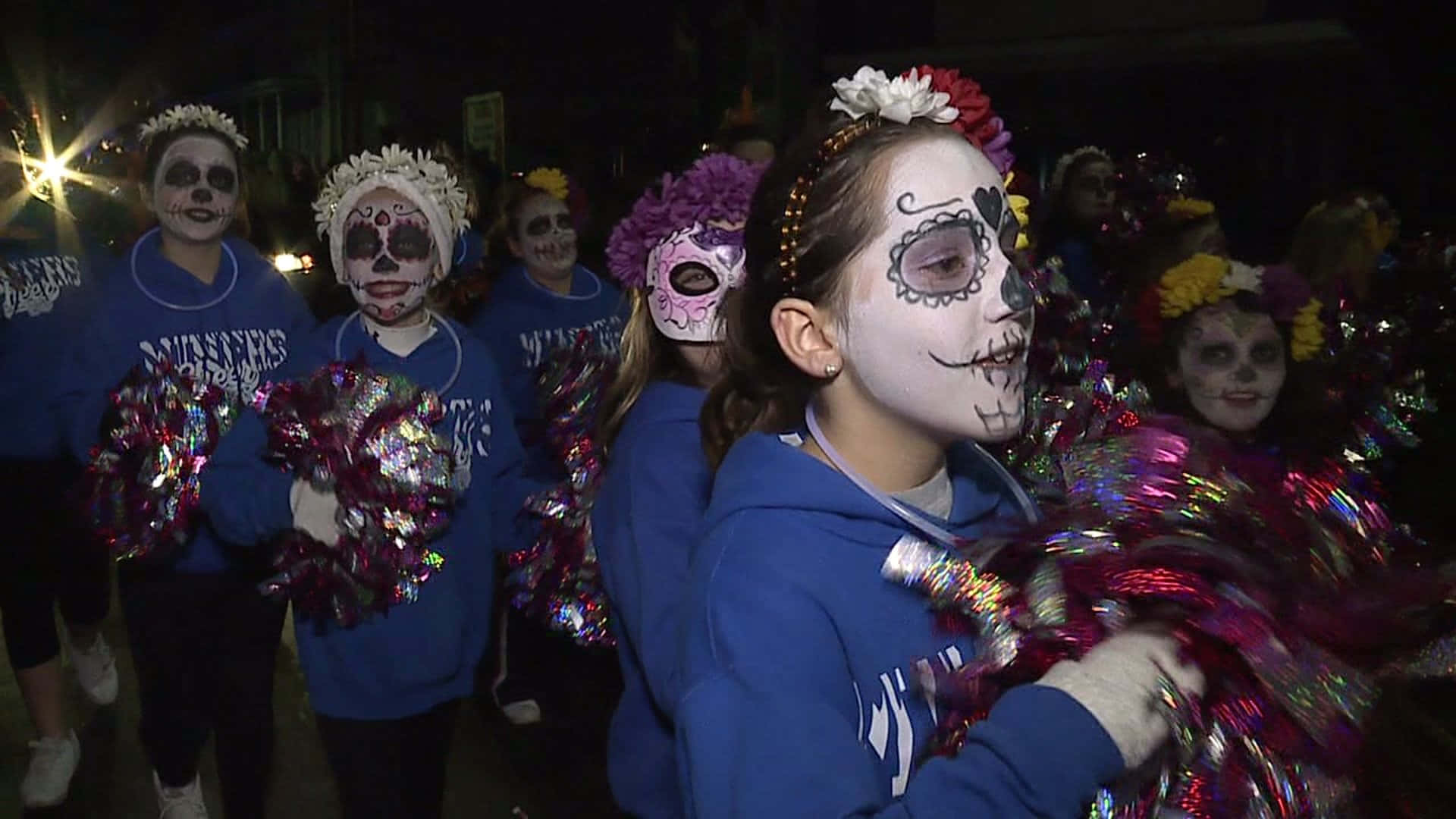 Halloween Parades 桌布 壁紙 — 年輕女孩穿著藍色連帽衫，帶有糖骷髏妝容和彩色絨球，在夜間節日遊行中行進。