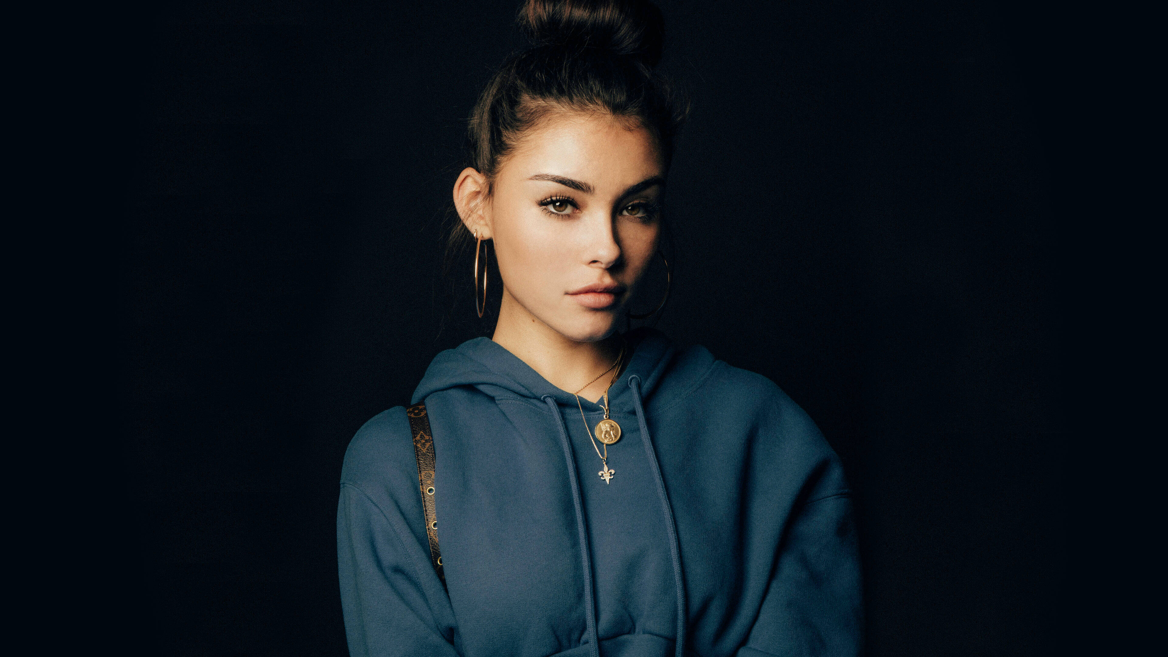 年輕女藝人 Madison Beer 壁紙 — 一位年輕女子將頭髮盤成髮髻，穿著藍色連帽衫和金色首飾，背景為深色。