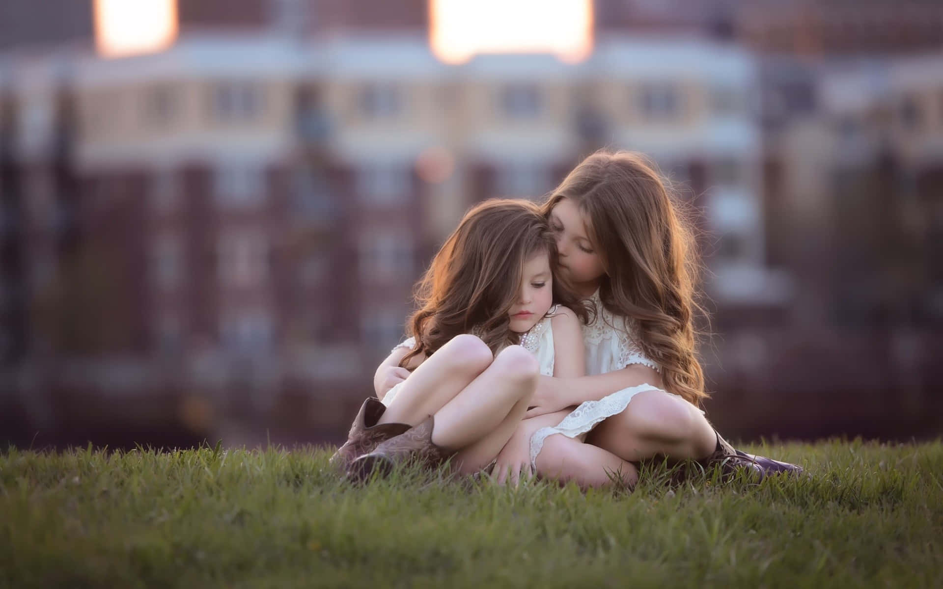 年輕姐妹圖 壁紙 — 兩個年輕女孩在草地上溫柔擁抱，金色夕陽灑在她們的頭髮和臉上。