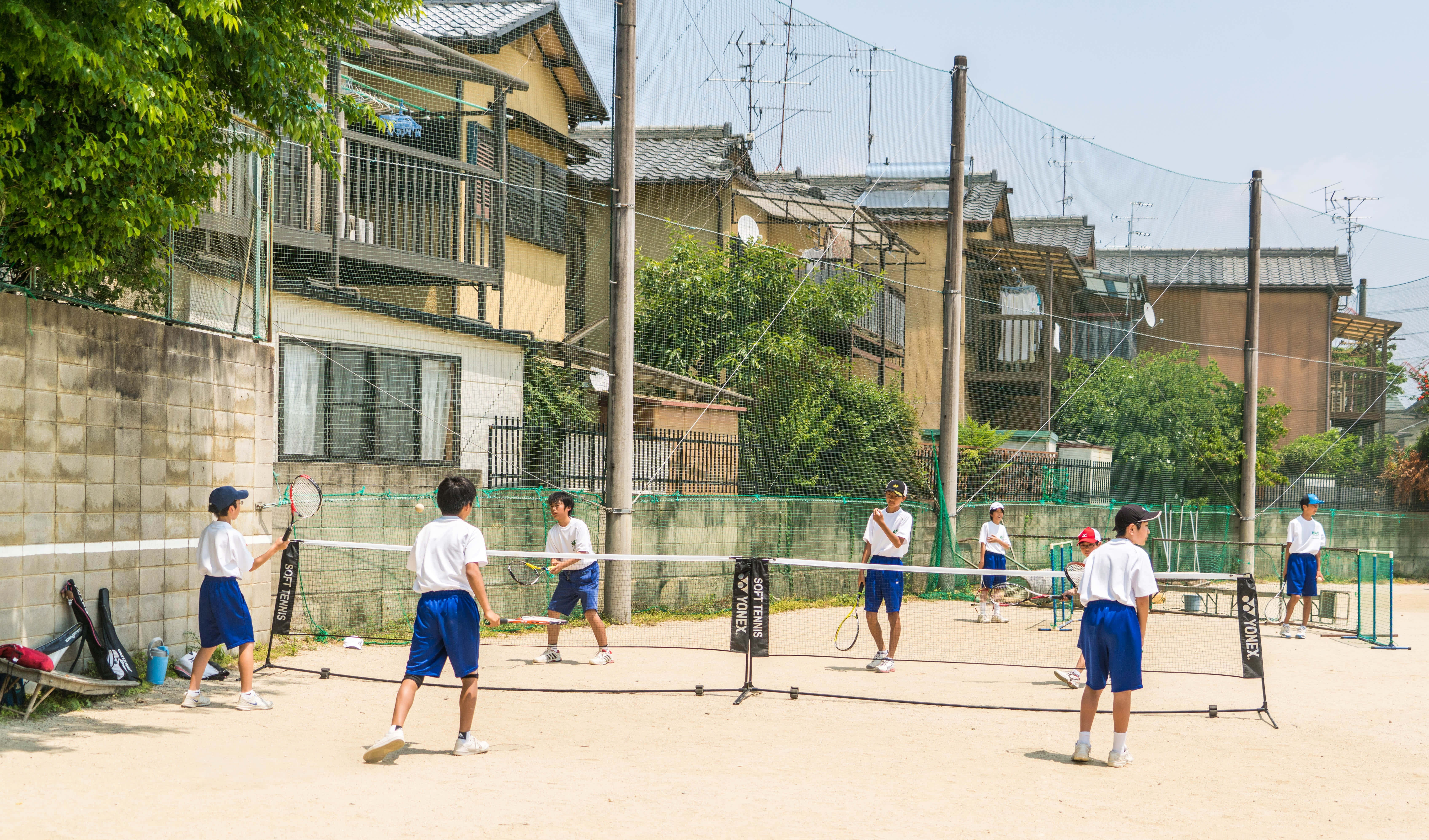 Badminton 桌布 壁紙 — 年輕學生穿著藍色短褲，在佈滿灰塵的球場上打網球，周圍是住宅房屋和綠樹，在明亮的日光下。