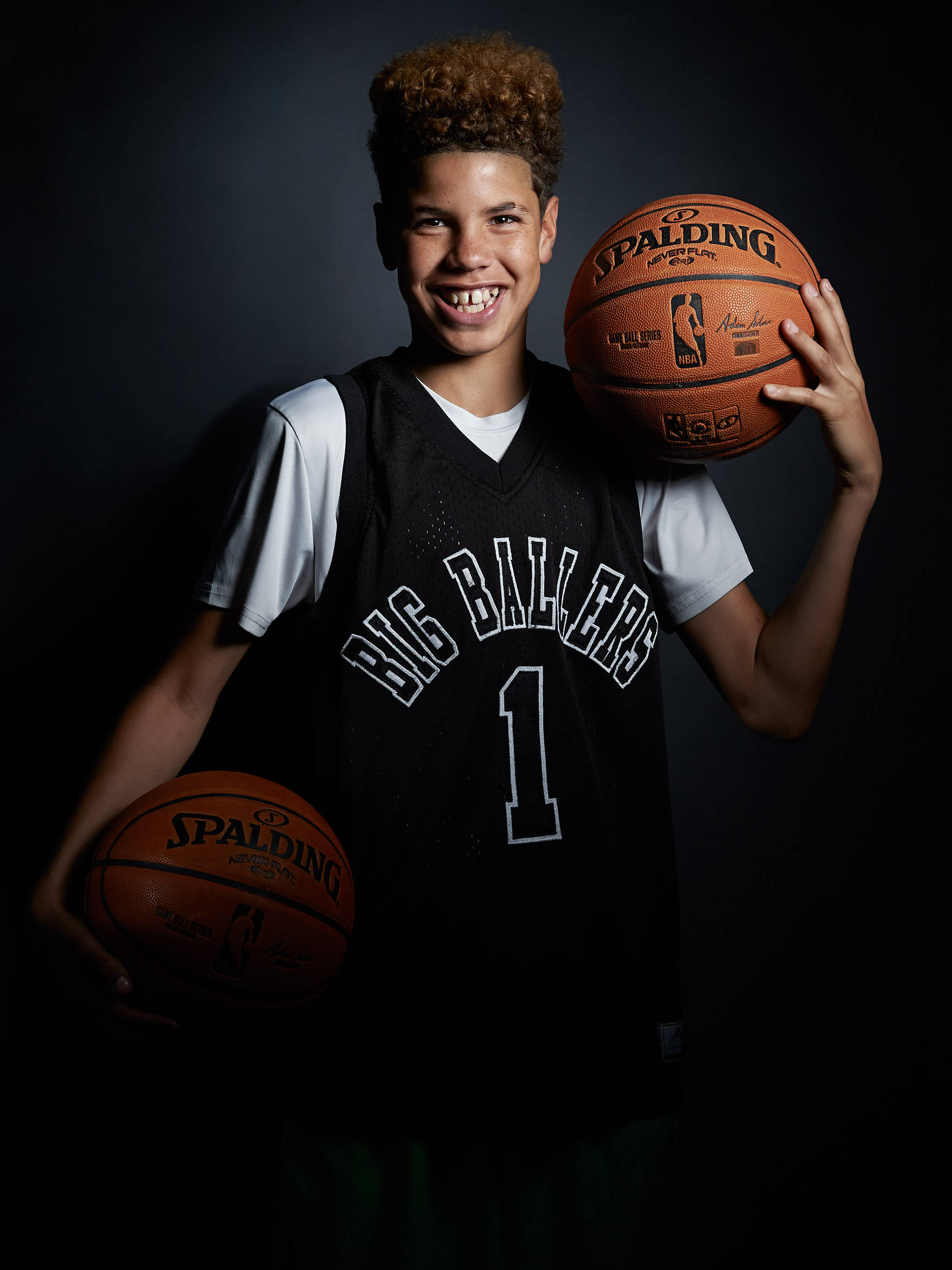 年輕拉梅洛·巴爾 壁紙 — 一位微笑的年輕籃球運動員身穿黑色 'Big Ballers' 球衣，在深色工作室背景下手持兩個 Spalding 籃球。