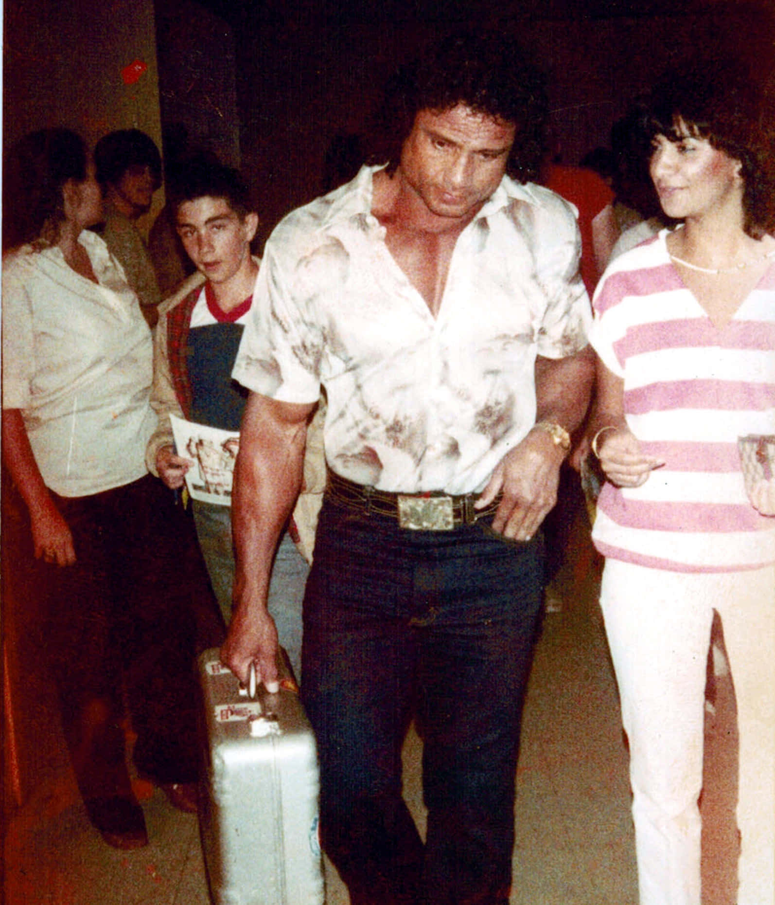 一個肌肉發達的男人帶著手提箱，穿著1980年代的服裝，穿過擁擠的室內派對。