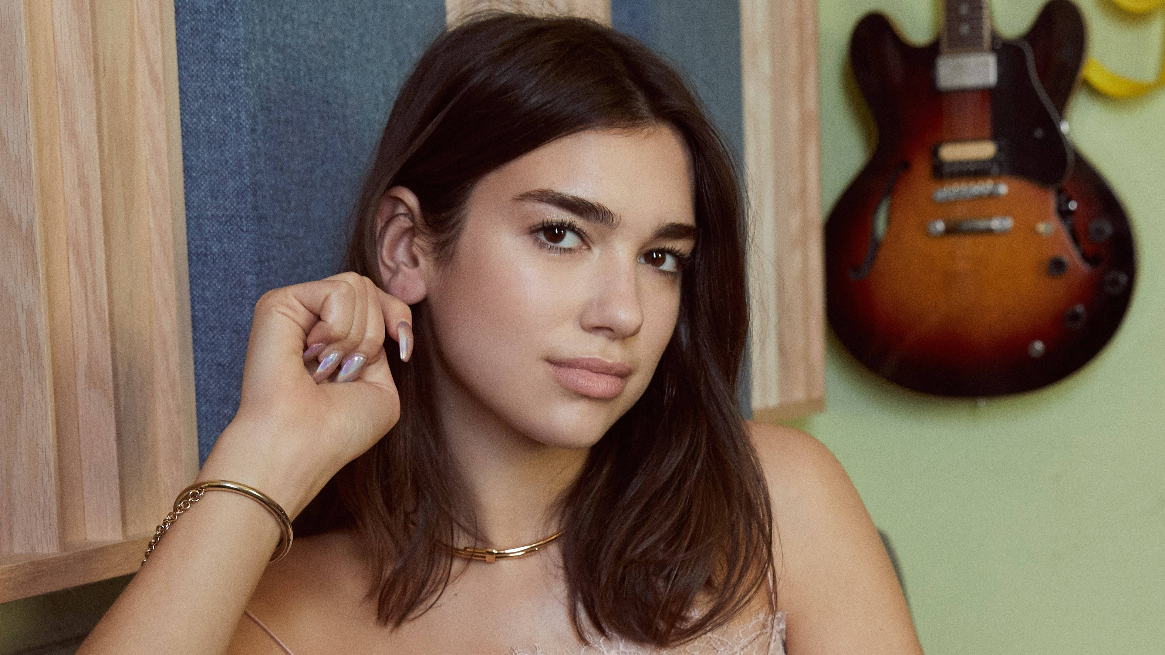 年輕時的 Dua Lipa 壁紙 — 一位黑髮戴金飾的年輕女子，在木鑲板和旭日紋結他的背景下，輕觸耳朵，溫柔凝視。