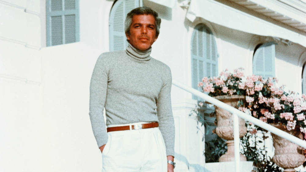 年輕時的曬黑 Ralph Lauren 壁紙 — 一個穿著灰色高領毛衣和白色褲子的男人自信地站在陽光普照的白色陽台上，旁邊是盛開的粉紅色花朵。
