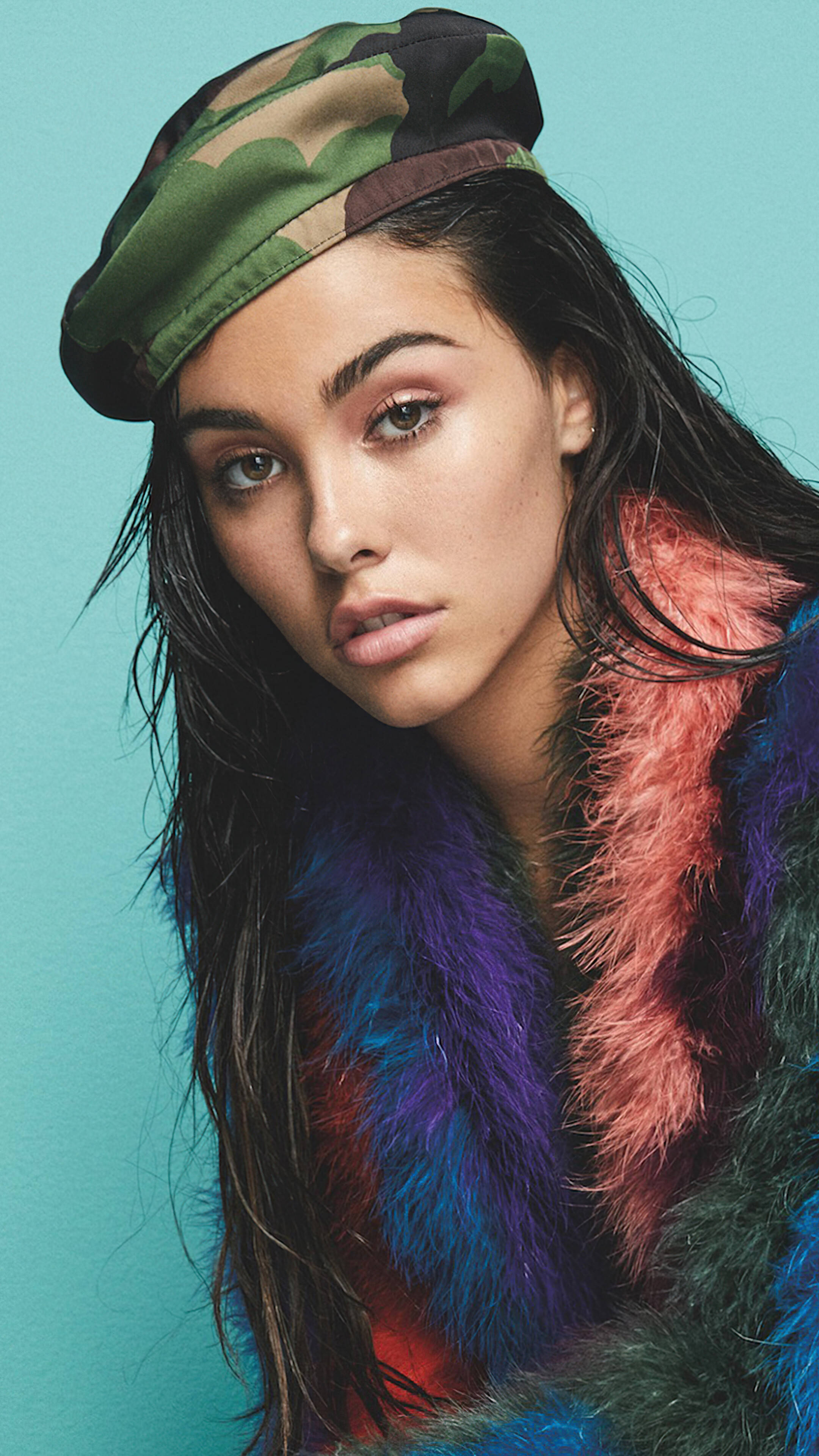 年輕歌手 Madison Beer 壁紙 — 一位深色頭髮的年輕女性戴著迷彩帽和鮮豔的多色毛領，背景為純青色。