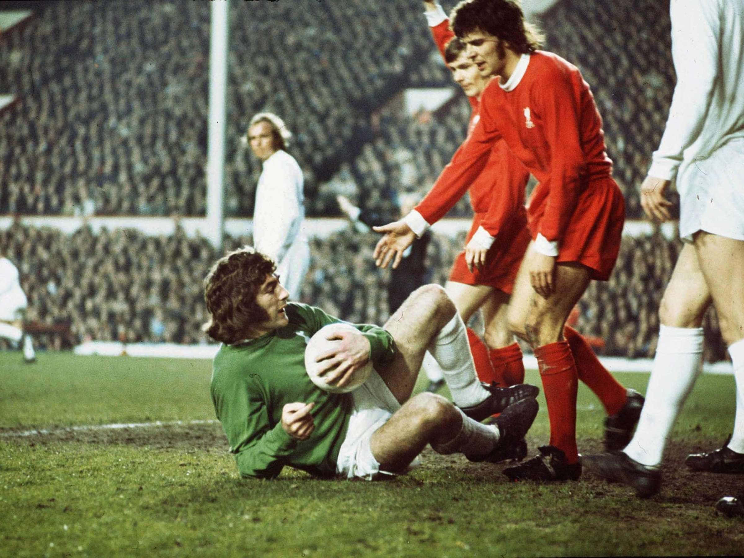 年輕球員 Pat Jennings 對陣 Peter Cormack 壁紙 — 一位身穿綠色球衣的復古足球員在草地上滑行時緊握著球，而身穿紅色球衣的對手則在附近虎視眈眈。