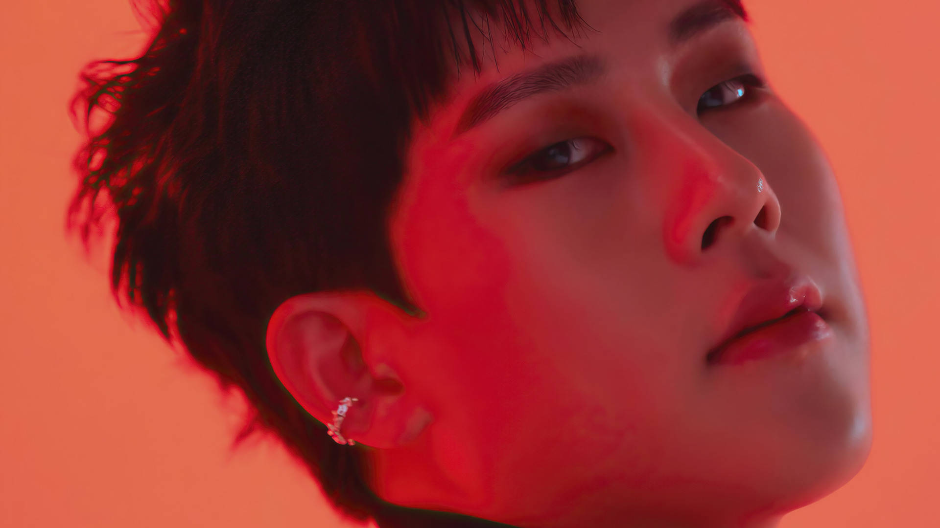 Monsta X Jooheon Fatal Love 壁紙 — 年輕男子近距離肖像，短黑髮，戴鑽石耳環，沐浴在強烈紅光中，背景為純橙色。