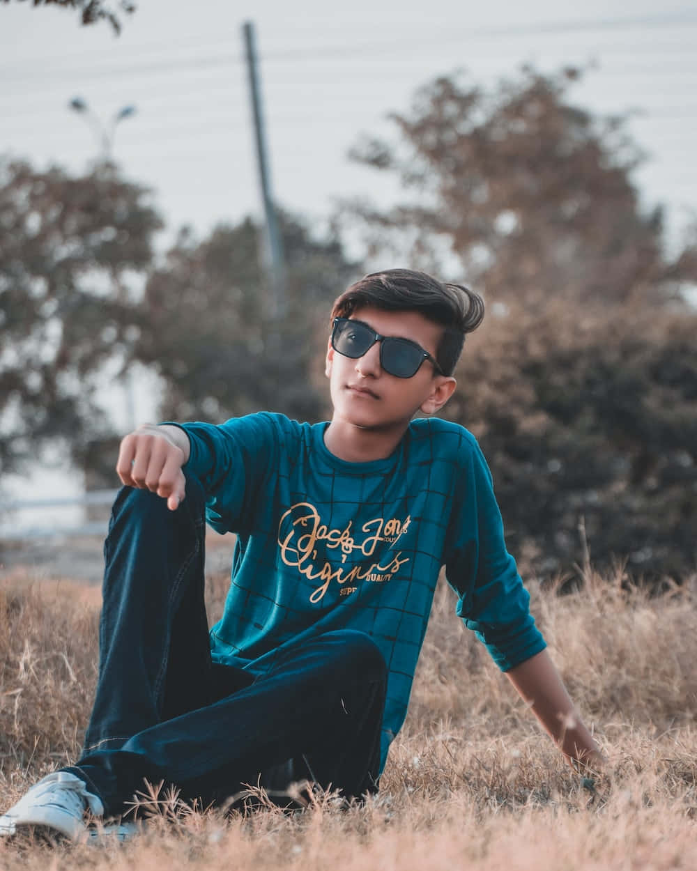 年輕男孩在都市中自信漫步 壁紙 — 一位年輕男子戴著太陽眼鏡，隨意地坐在乾草上，背景有樹木和電線，畫面柔和。