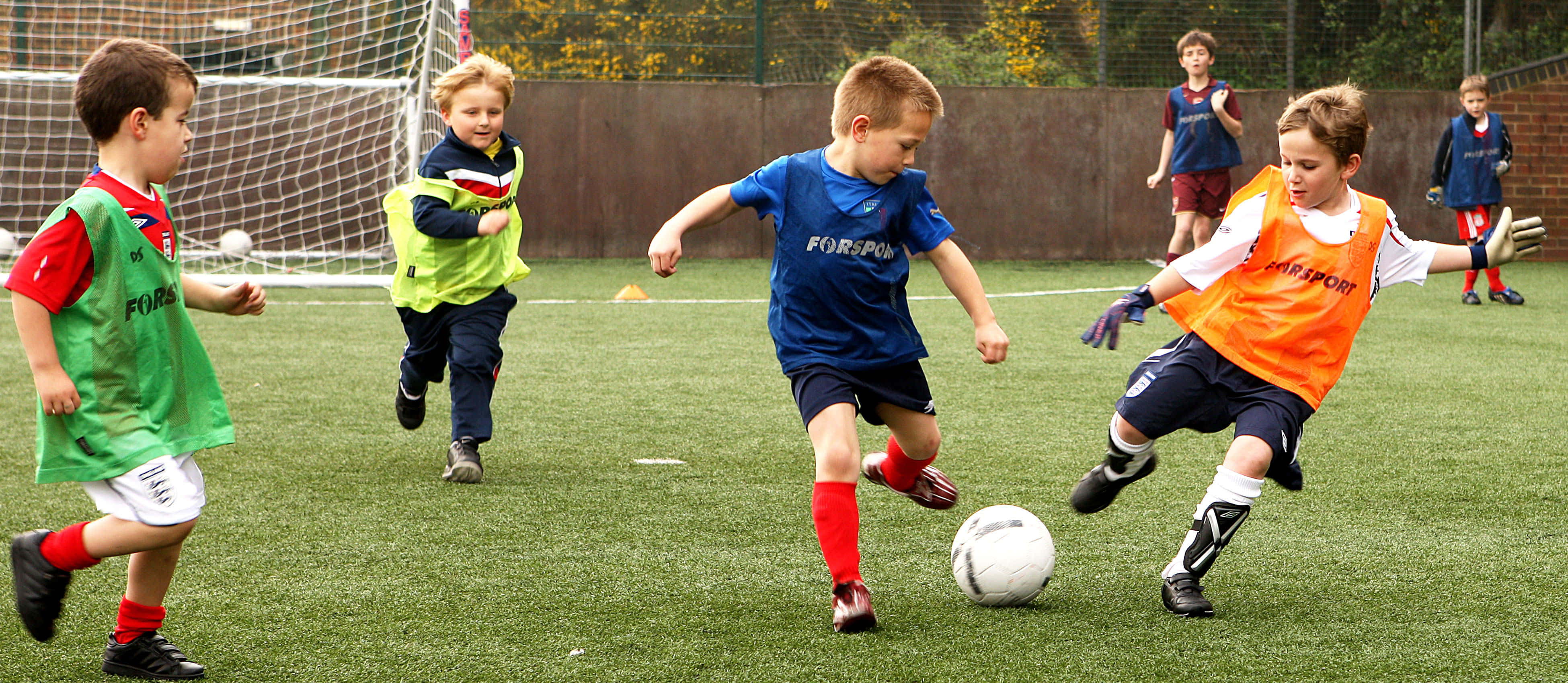 Kids Soccer 桌布 壁紙 — 年輕男孩穿著色彩繽紛的訓練背心，在充滿活力的綠色人造草地球場上精力充沛地爭奪足球。