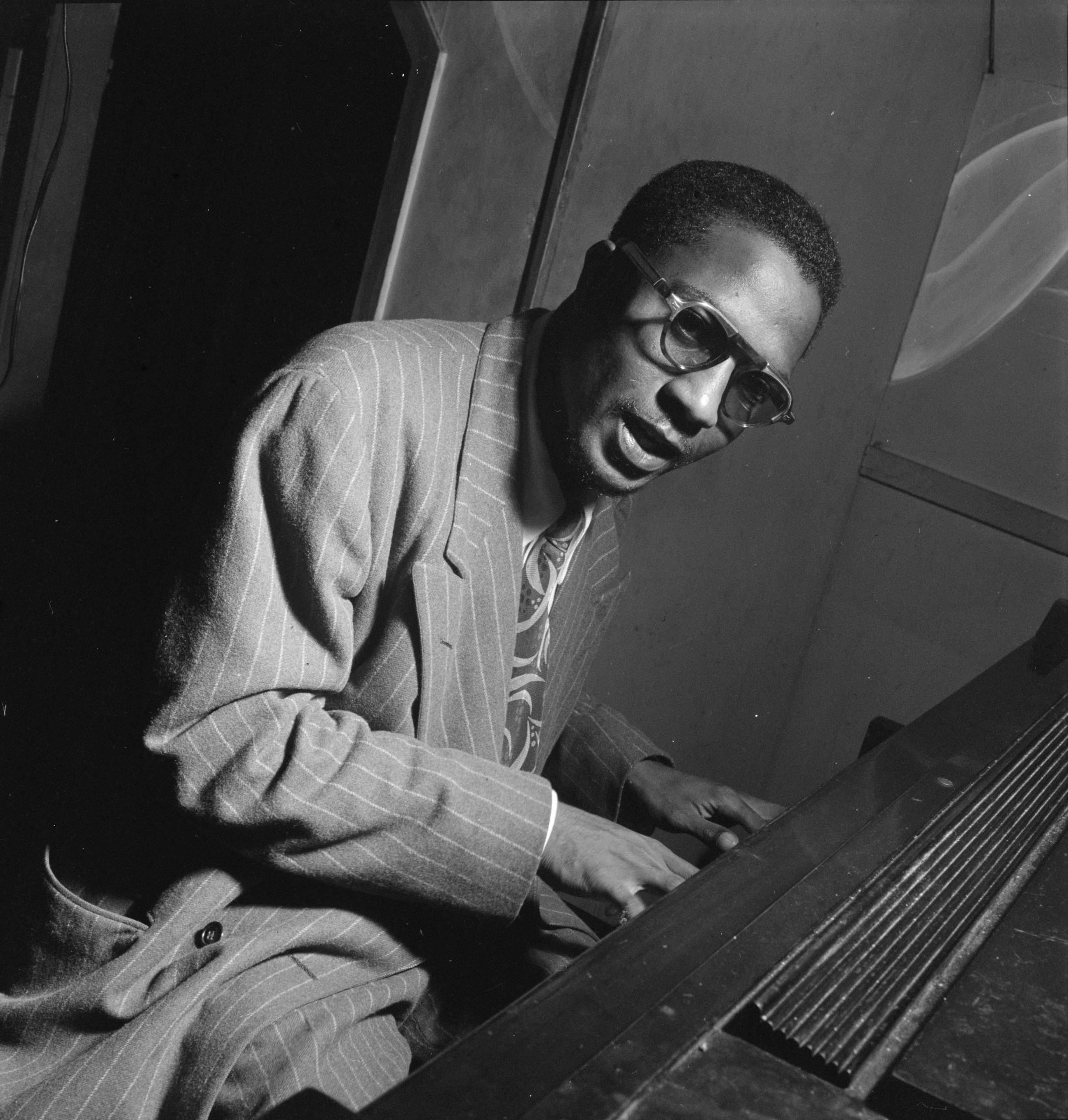 年輕的 Thelonious Monk 演奏鋼琴 壁紙 — 一名身穿細條紋西裝、戴眼鏡的男子，在昏暗的房間中熱情地彈奏鋼琴，以引人注目的黑白影像捕捉。