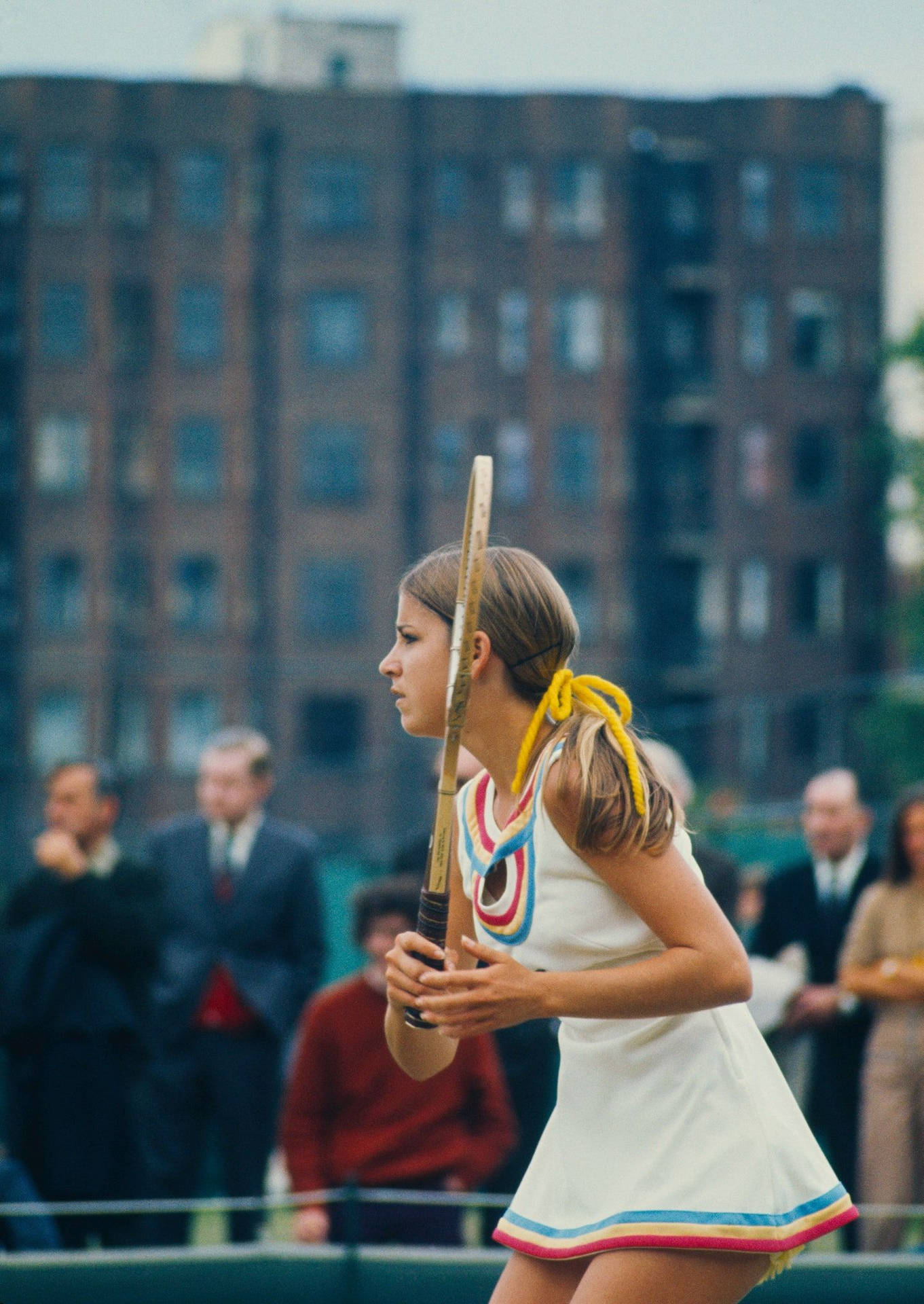 1970年代服裝的女網球運動員在球場上拿著球拍，後面有磚砌建築。