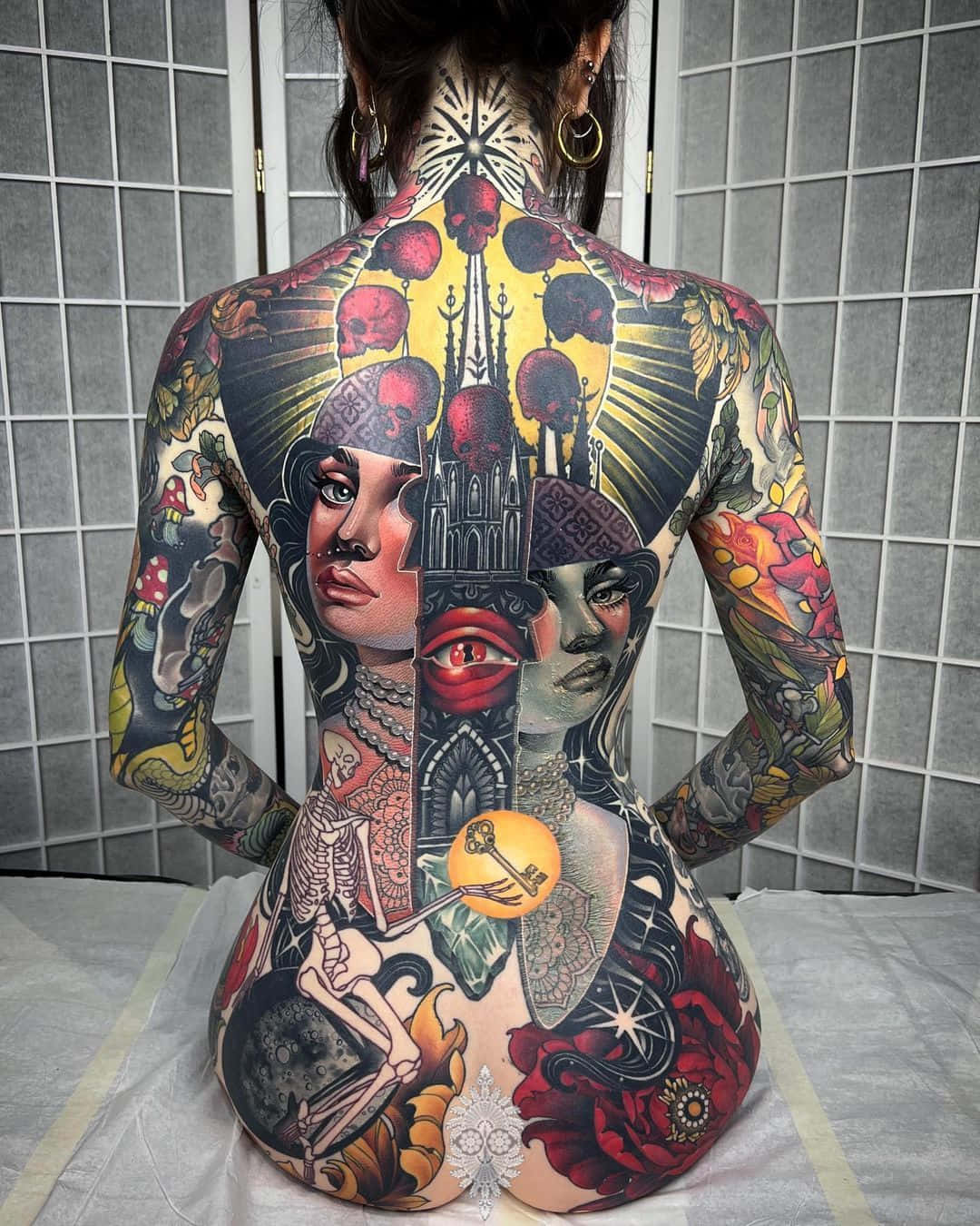 年輕紋身女孩展示她的藝術創作 壁紙 — 一個人的背部佈滿精細多彩的紋身，圖案包括骷髏頭、玫瑰、眼睛和哥德式建築，背景是瓷磚牆。