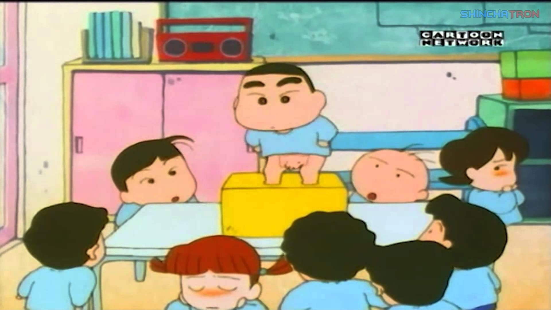 年輕蠟筆小新在校園 壁紙 — 一個卡通男孩站在課室裏的黃色箱子上，周圍穿着藍色校服的同學們抬頭看着他。