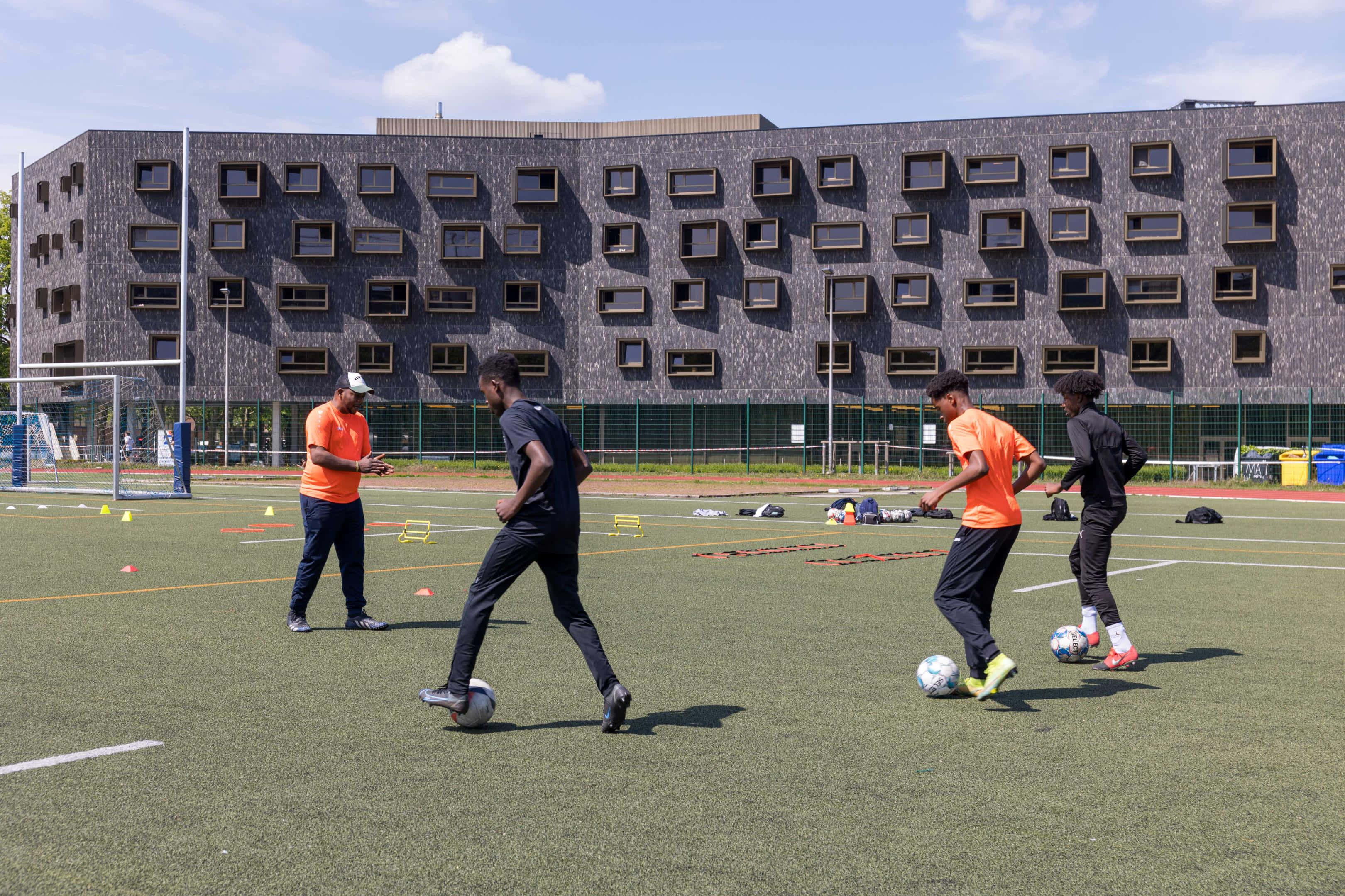 sport 桌布 壁紙 — 年輕足球運動員在明亮的日光下於綠色草地場上運球，背景是一座現代建築。