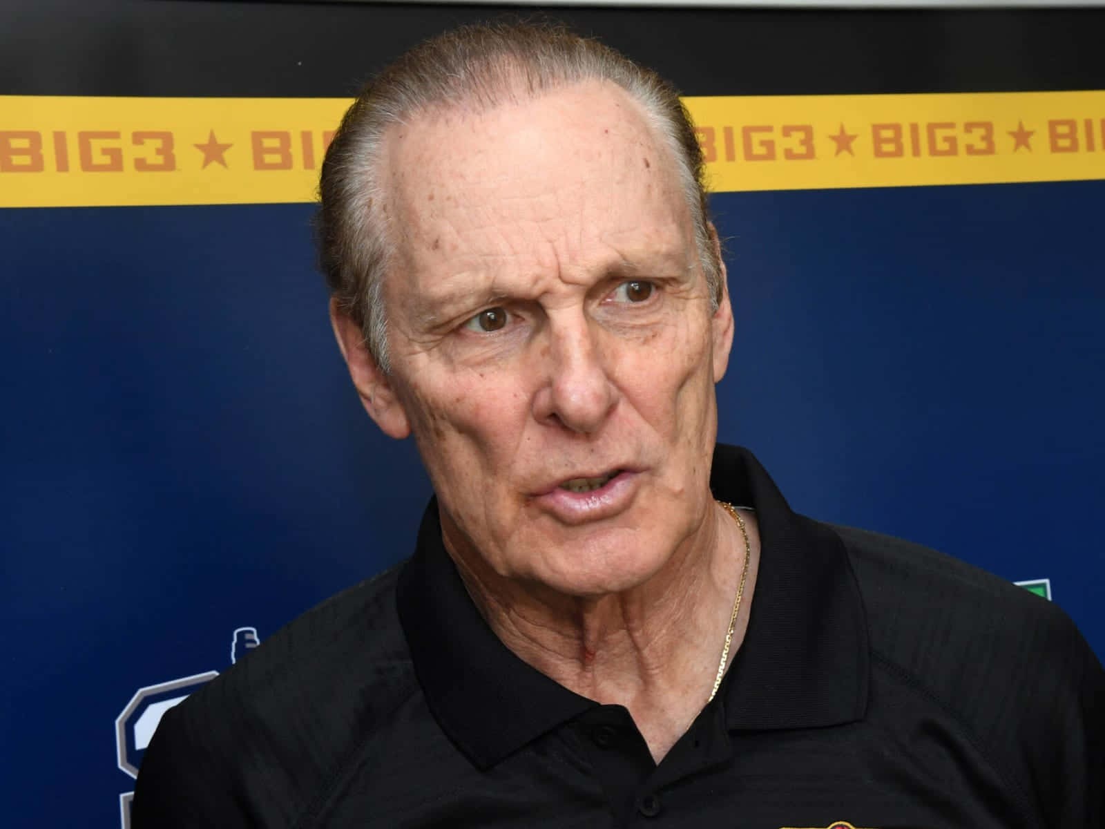 年長的NBA瑞克·巴里 壁紙 — 一位年長男士的特寫肖像，眼神銳利，身穿黑色Polo衫，背景為藍色並帶有重複的黃色BIG3標誌。