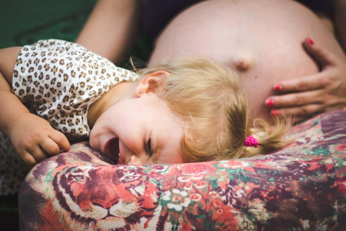 幼童在孕婦肚子上哭泣 壁紙 — 一個穿著豹紋襯衫的小女孩，將頭靠在老虎圖案的枕頭上，在孕婦的肚子旁邊哭泣。