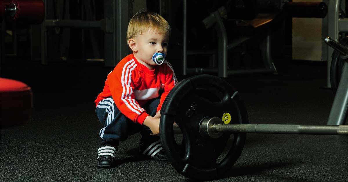 穿著紅色運動服的幼兒在健身房裡含著奶嘴拿著槓鈴。