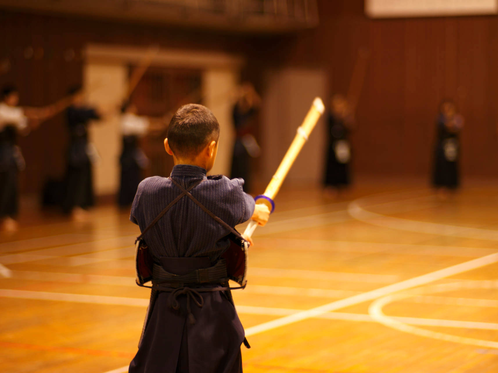 幼童學習劍道武術 壁紙 — 一個身穿傳統武術裝備的小男孩在燈光溫暖的體育館裡用木劍練習。