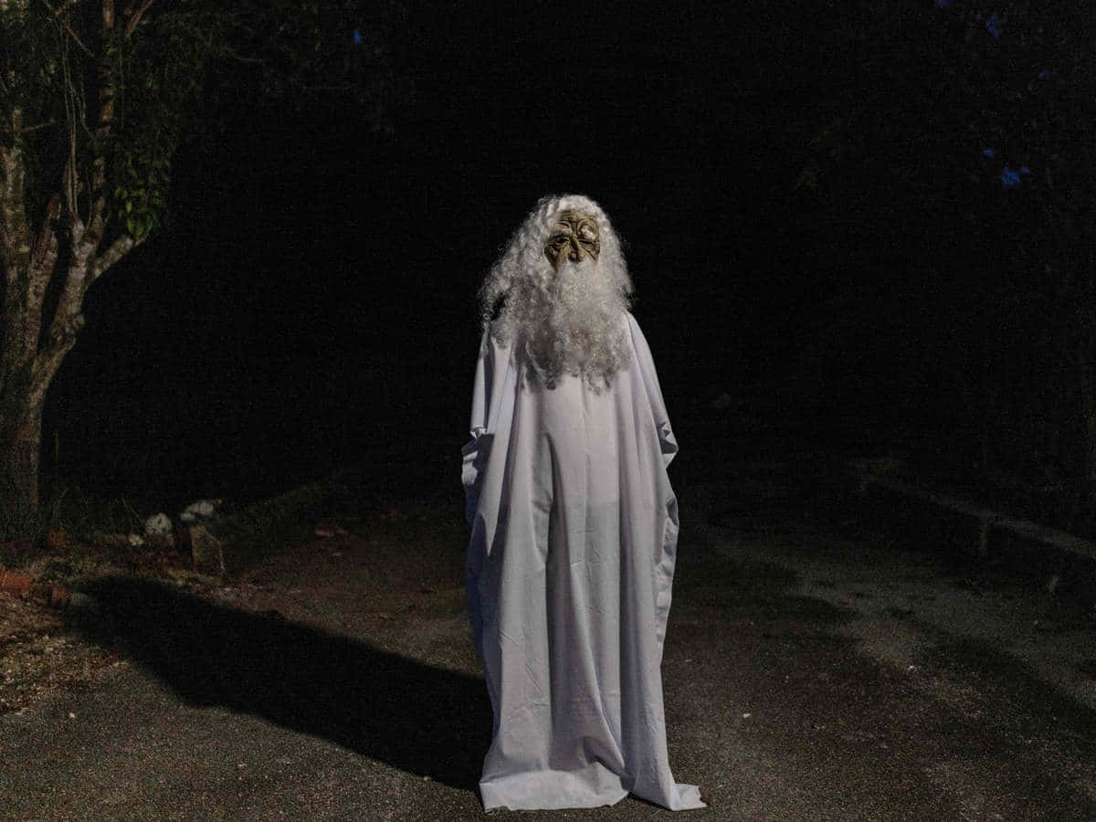 一個長著白色長髮和鬍鬚的幽靈般的身影，夜晚獨自站在黑暗的森林小徑上。