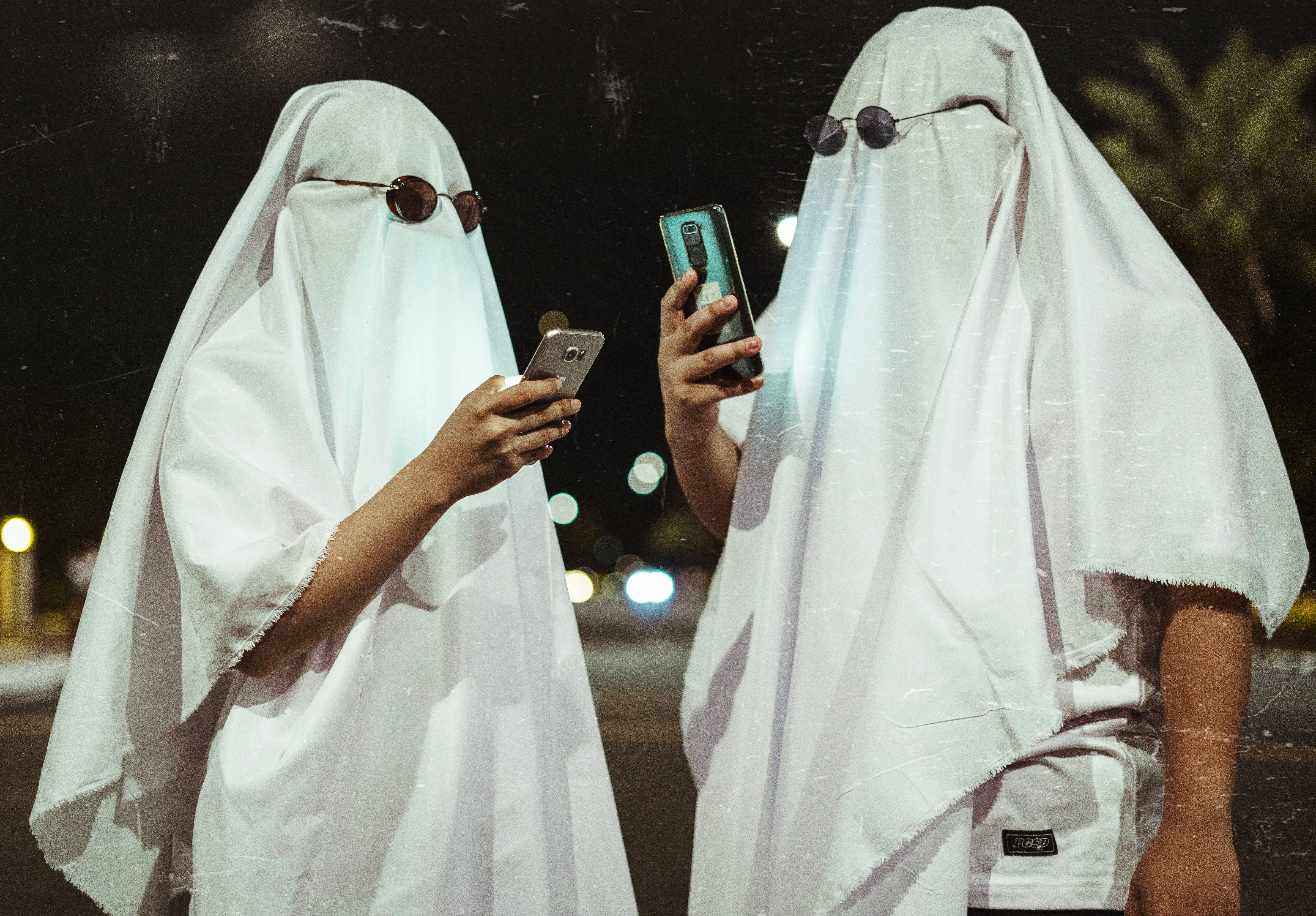 幽靈萬聖節服裝電腦壁紙 壁紙 — 兩個穿著白色鬼魂服裝、戴著太陽眼鏡的人物在夜晚站立，全神貫注於他們發光的智能手機，背景是模糊的城市景觀。