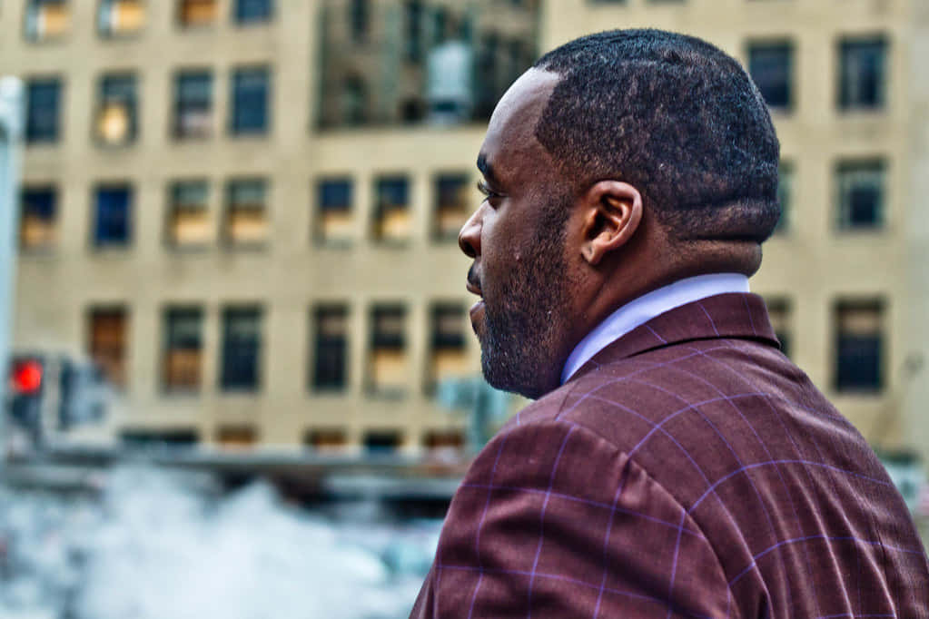 底特律前市長Kwame Kilpatrick肖像 壁紙 — 一名身穿栗色格紋西裝的男子側面站立，背景是模糊的城市景觀，窗戶透出溫暖燈光。