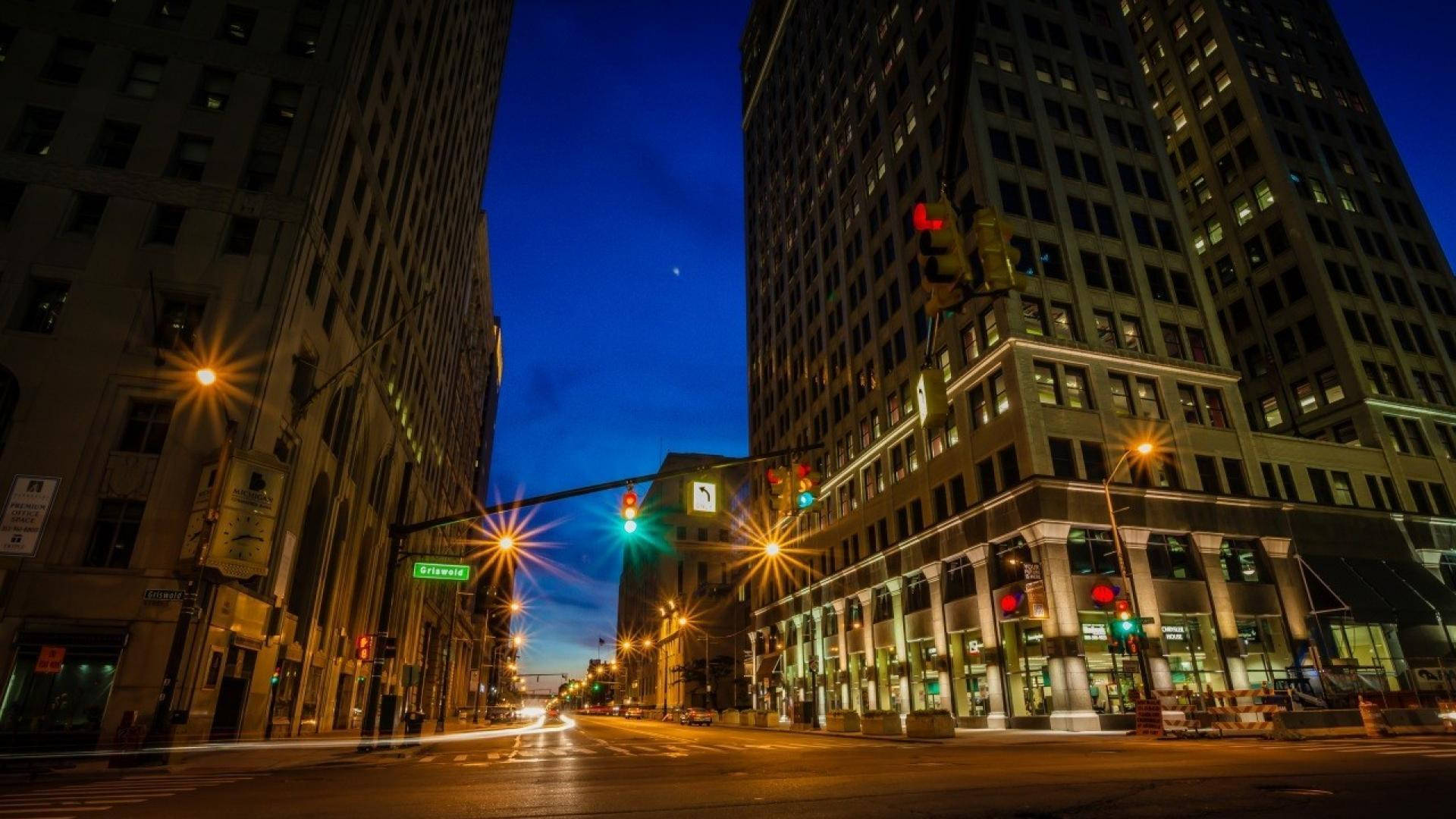 底特律夜間街景 壁紙 — 一張長時間曝光的城市街道照片，在暮色中，高聳建築物之間有發光的交通燈和汽車燈光軌跡。