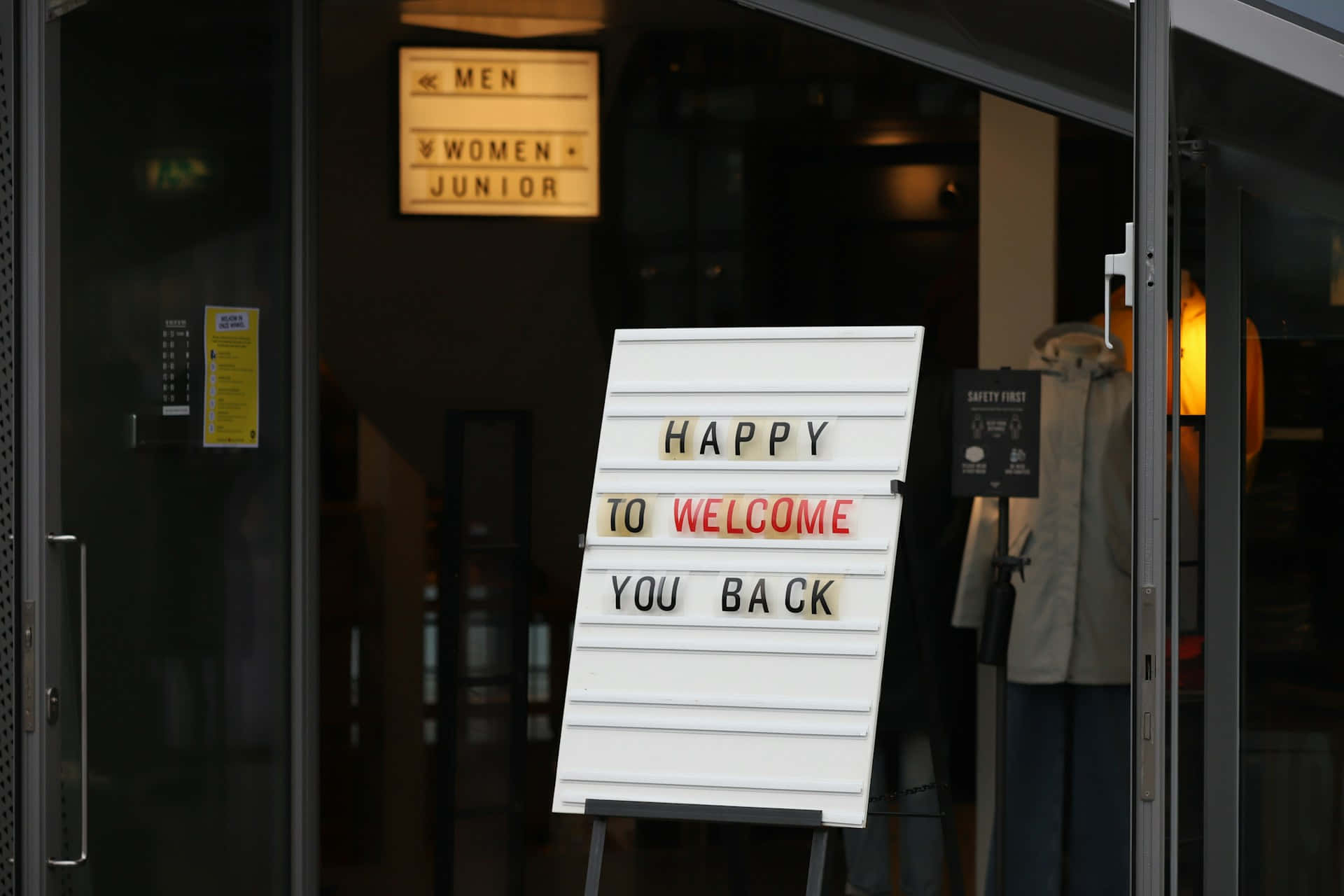 店外歡迎回來標誌 壁紙 — 一個白色燈箱標牌，上面寫著「HAPPY TO WELCOME YOU BACK」，豎立在帶有玻璃門的商店入口外，上方有一個發光的「MEN WOMEN JUNIOR」標牌。