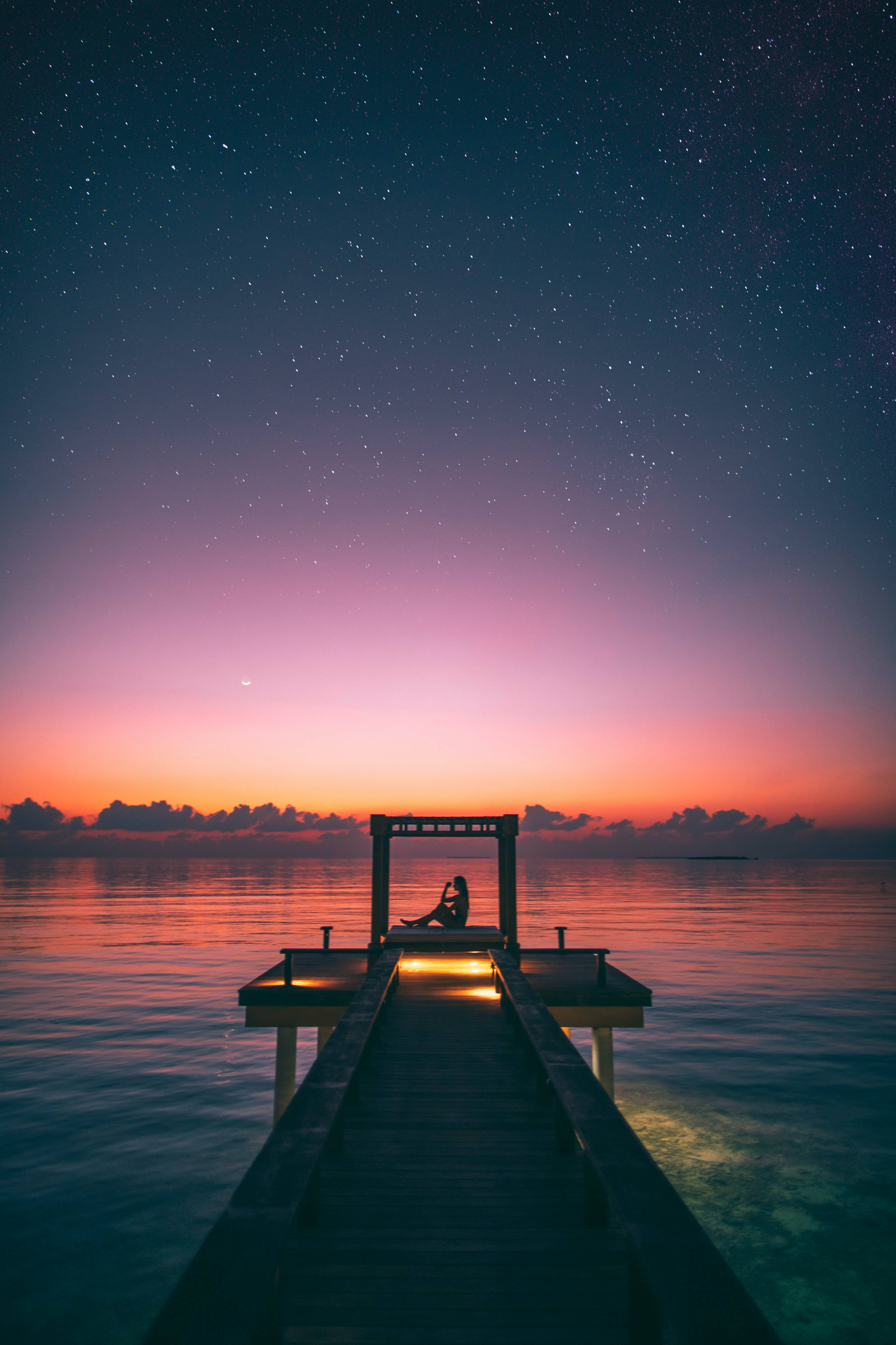 度假碼頭夕陽 壁紙 — 一個孤獨的身影坐在木碼頭上，在星空暮色天空下，粉紅、橙色和深藍色的漸變映照在平靜的水面上。