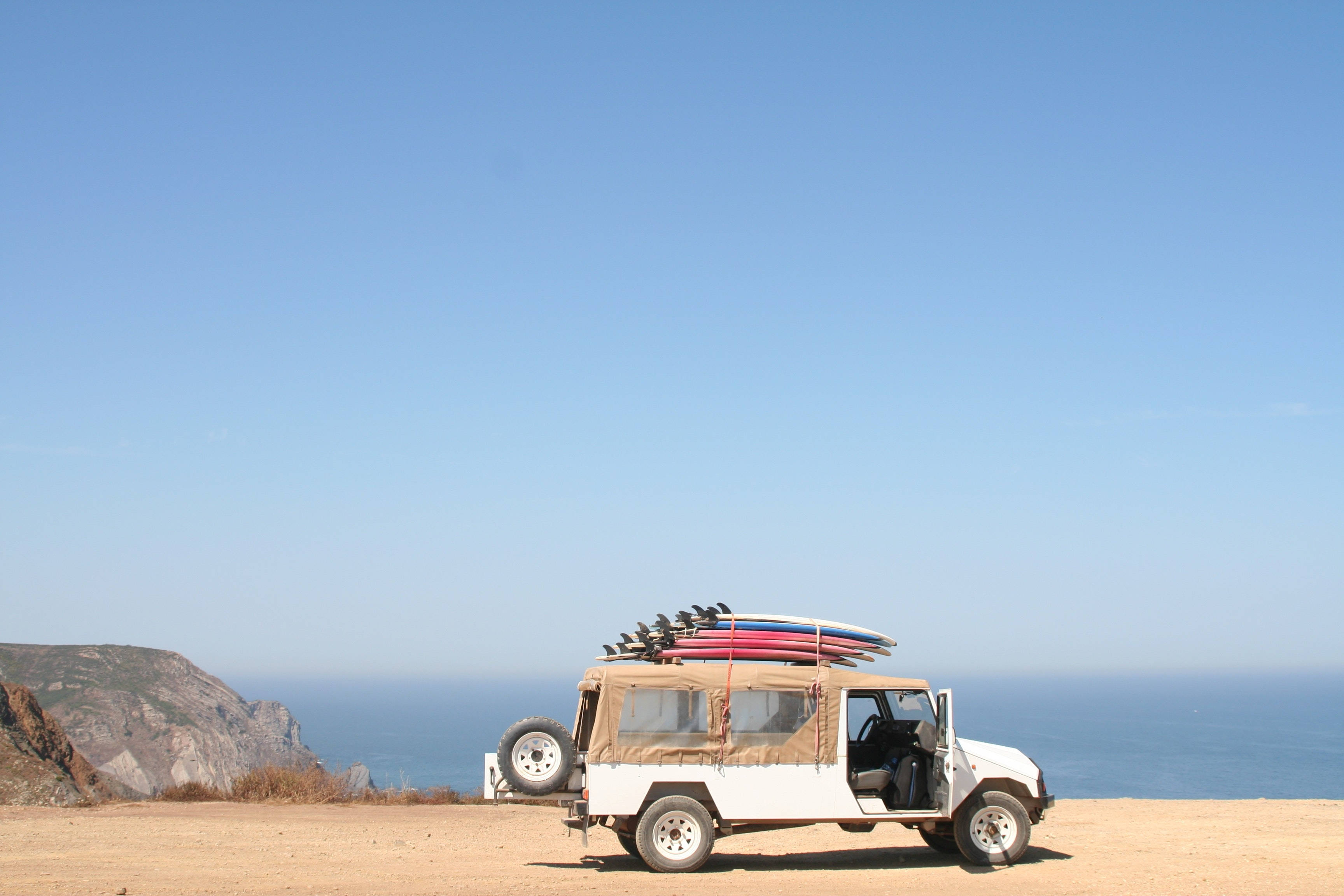 度假衝浪板吉普車 壁紙 — 一輛白色 safari 吉普車，車頂綁著衝浪板，停在陽光明媚的懸崖上，俯瞰著清澈藍天下的海洋。