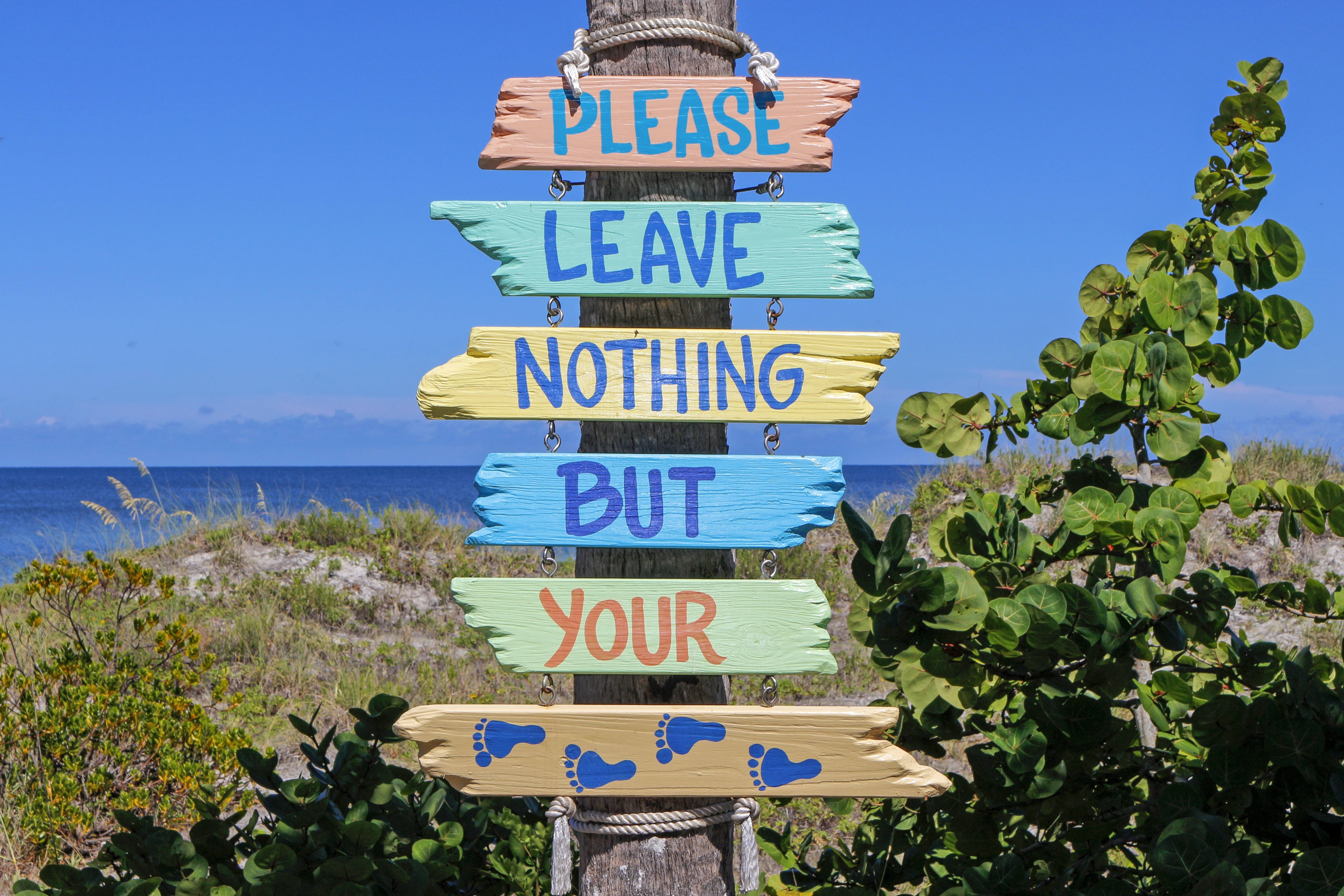 度假靈感語錄 壁紙 — 色彩繽紛的木製標牌上寫著「PLEASE LEAVE NOTHING BUT YOUR FOOTPRINTS」，豎立在沙灘柱上，背景是清澈的藍天和海洋。