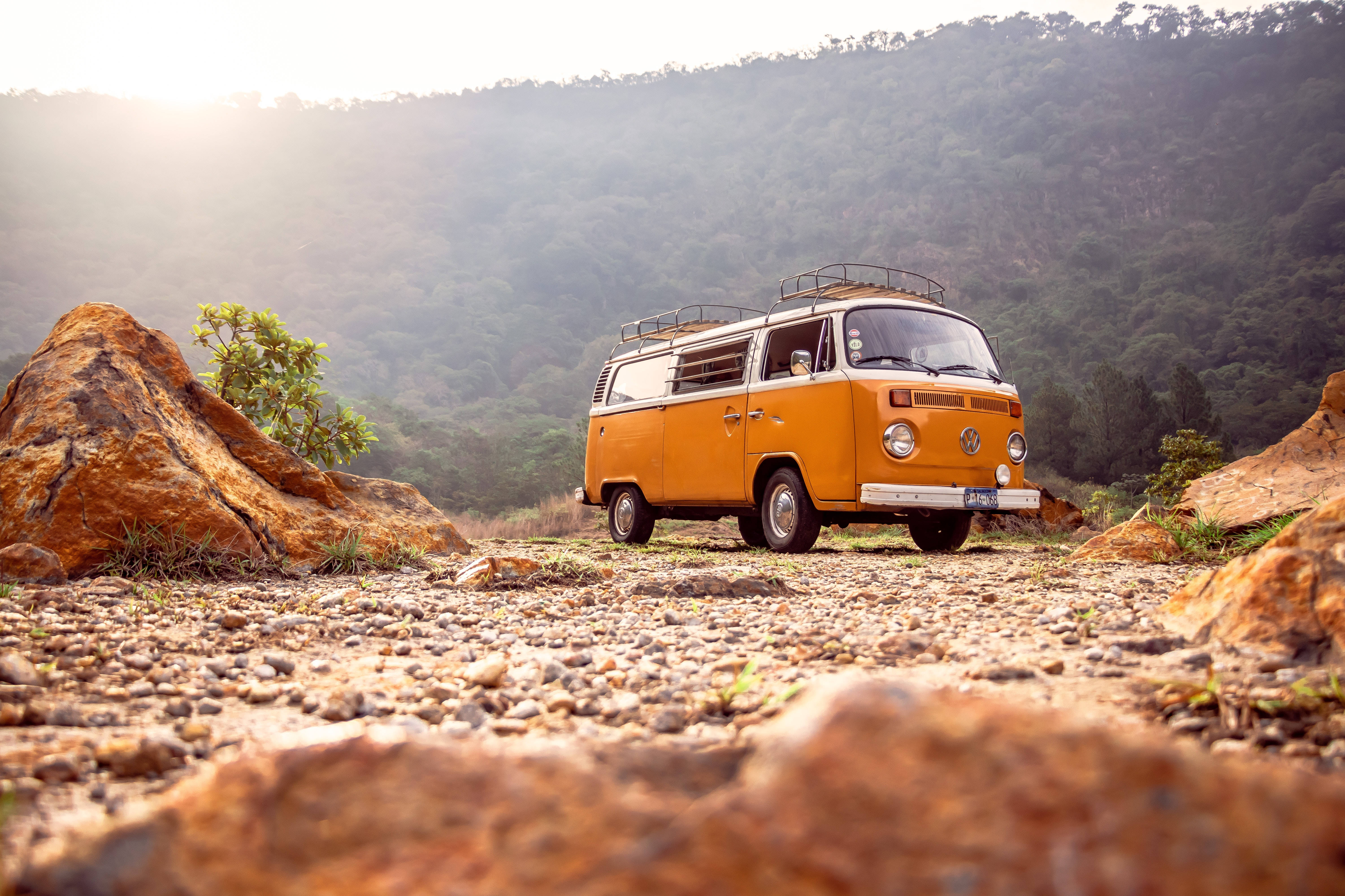 度假黃色小巴 壁紙 — 一輛復古橙色福士偉根露營車停泊在岩石小徑上，背景是陽光普照的山丘和樹木。