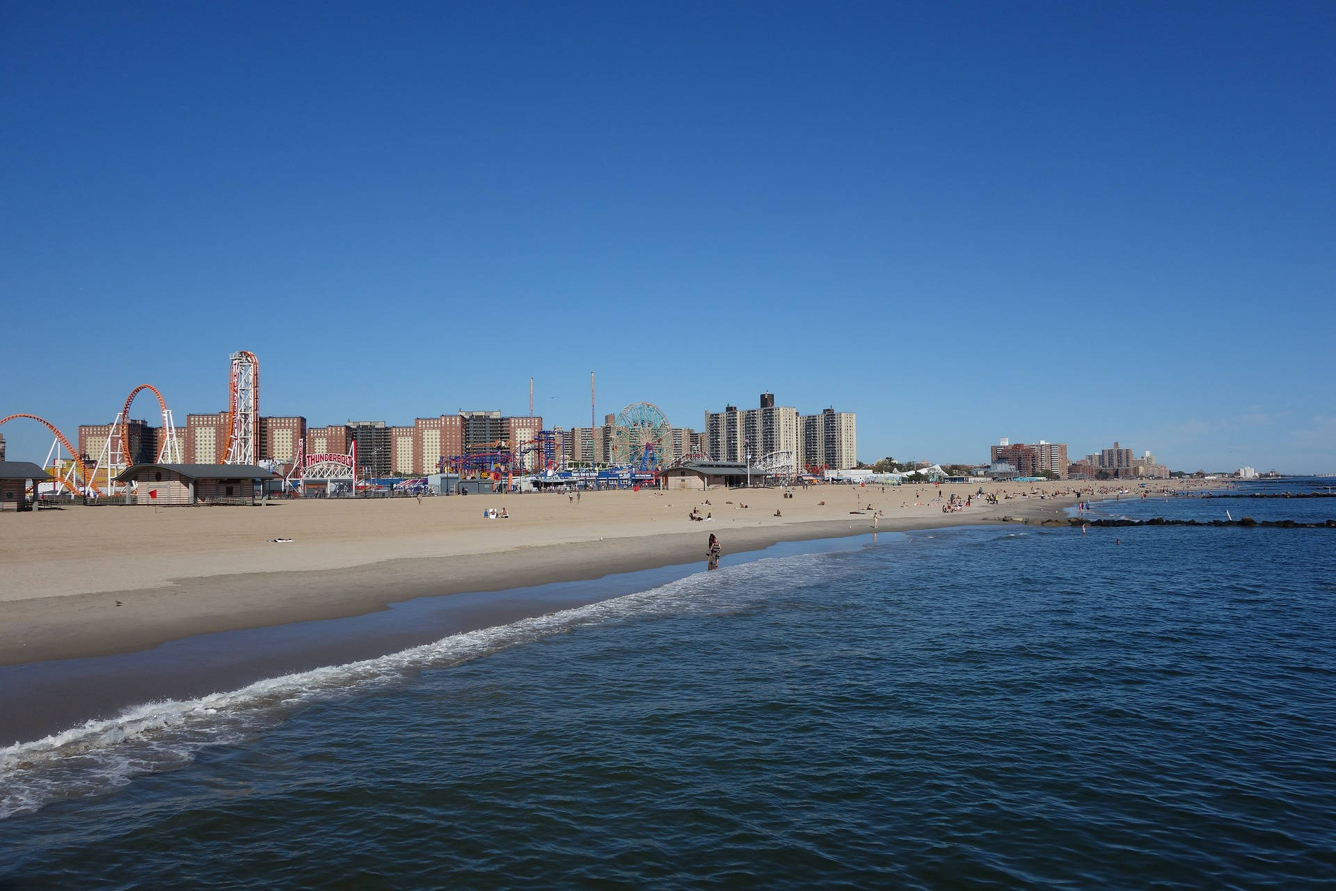 海浪拍打沙灘，背景有遊樂園和建築物，天空清澈湛藍。