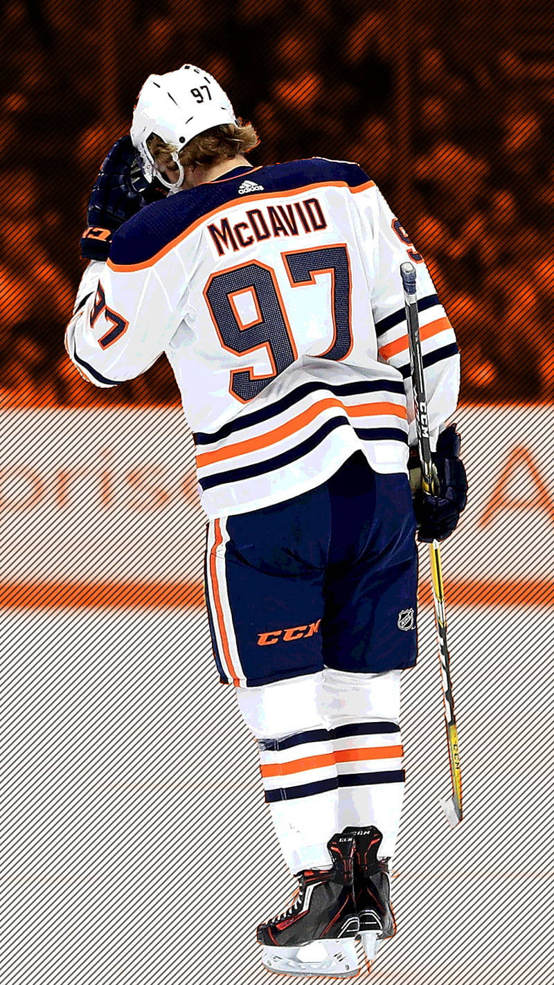 康納·麥克戴維 球衣編號97 埃德蒙頓油人隊 壁紙 — 冰球運動員 Connor McDavid 身穿白色、橙色和藍色 97 號球衣，站在冰上手持球棍，背景人群模糊。